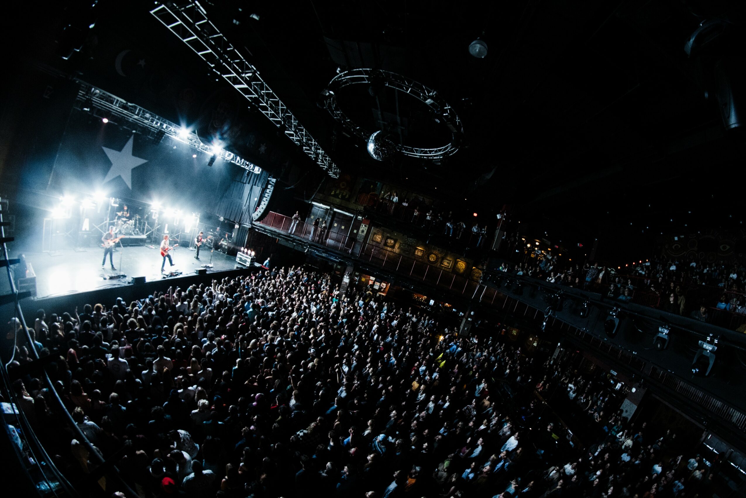 Concert stage with bright lights, performers, and a large audience in a dark indoor venue.