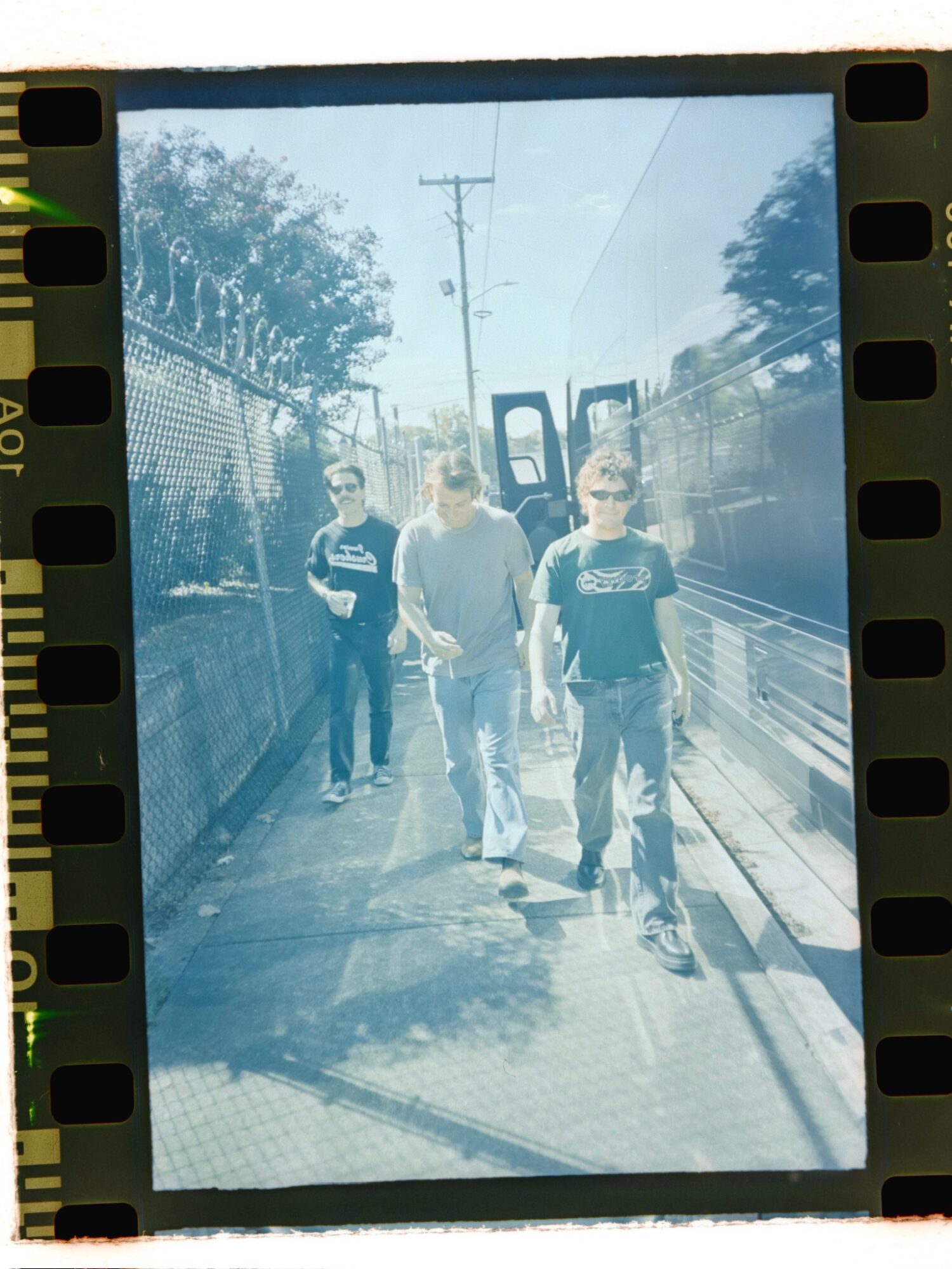Three people walking on a sidewalk with trees and a fence, one person wearing sunglasses, sunny day.