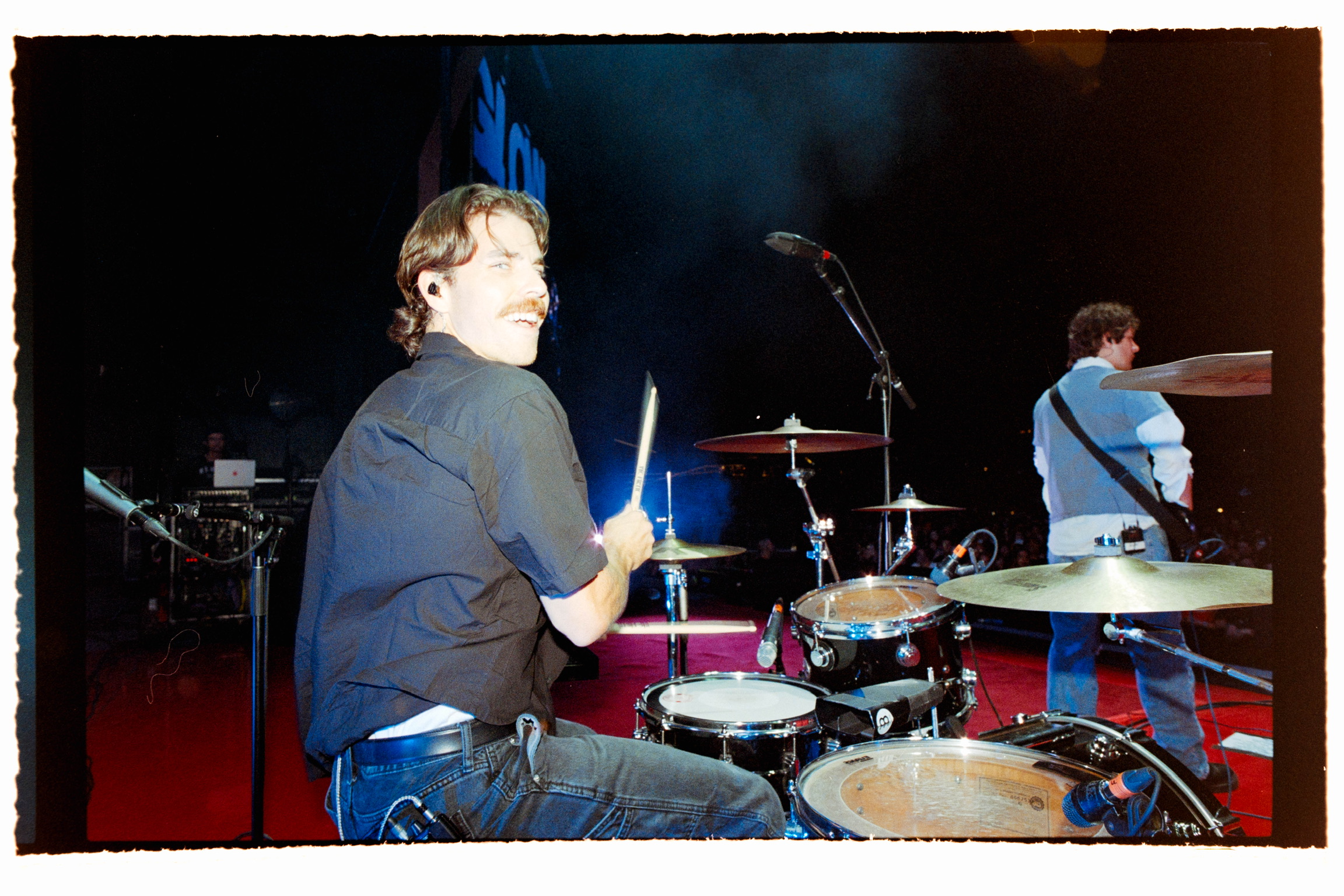Person playing drums on stage with another person in the background, stage lighting visible.