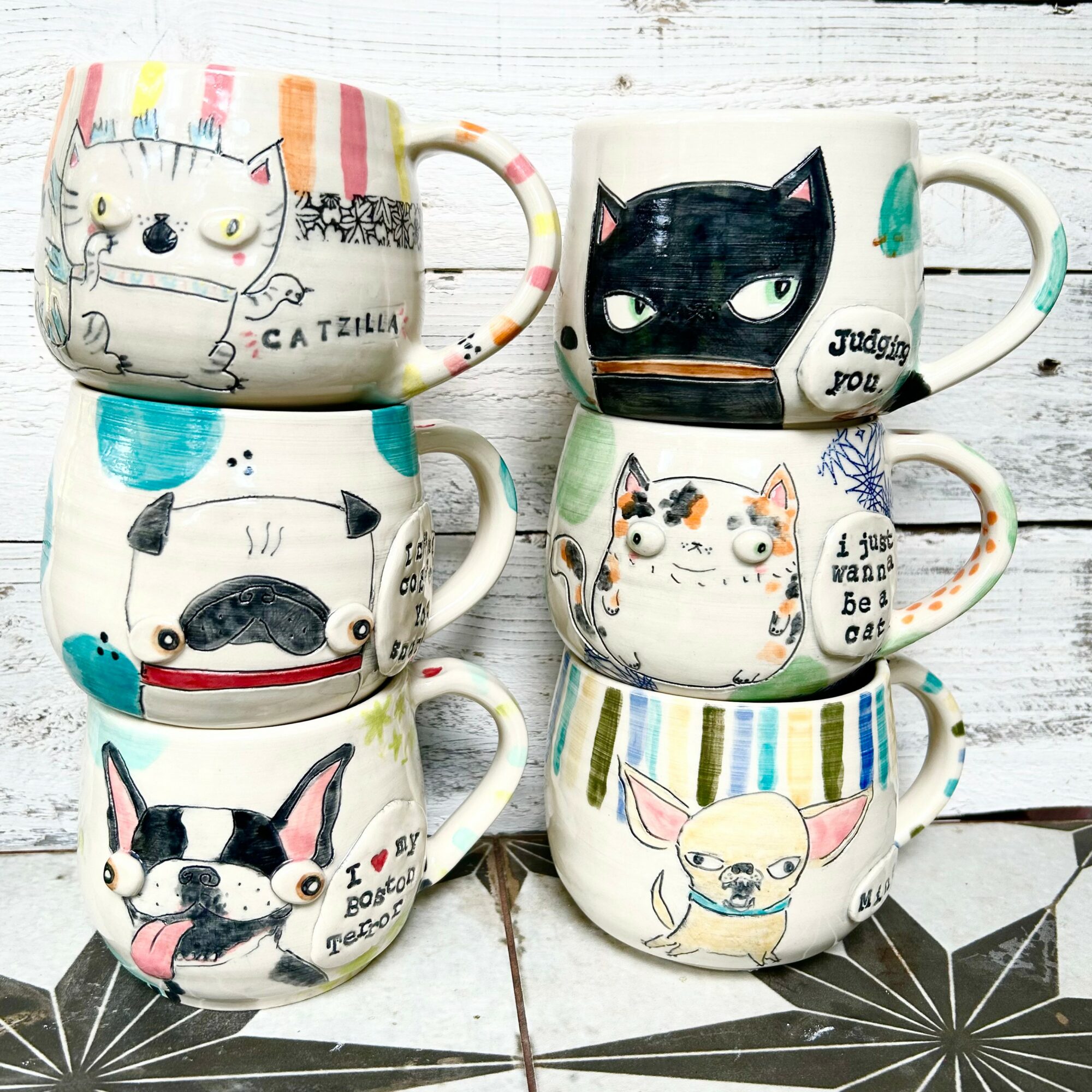 Six decorated mugs with various cat illustrations on a white wooden surface.