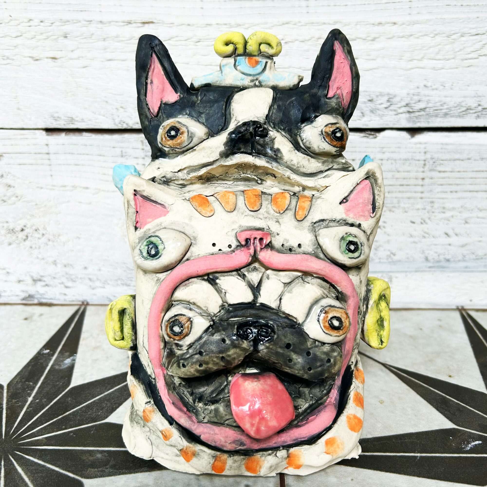 Colorful ceramic dog face sculpture with multiple faces, set against a white wooden background and black patterned surface.