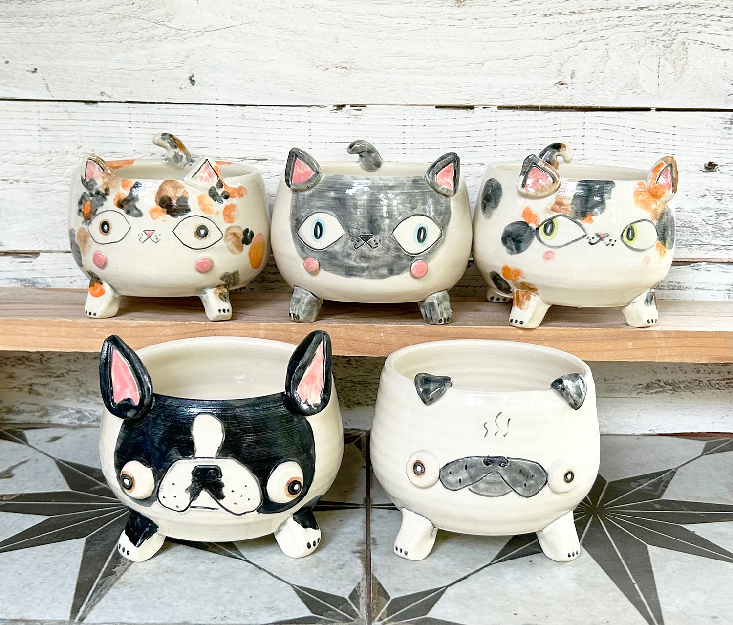 Five animal-shaped ceramic planters with faces, arranged on a wooden shelf and tiled floor, featuring cats and dogs with different expressions.