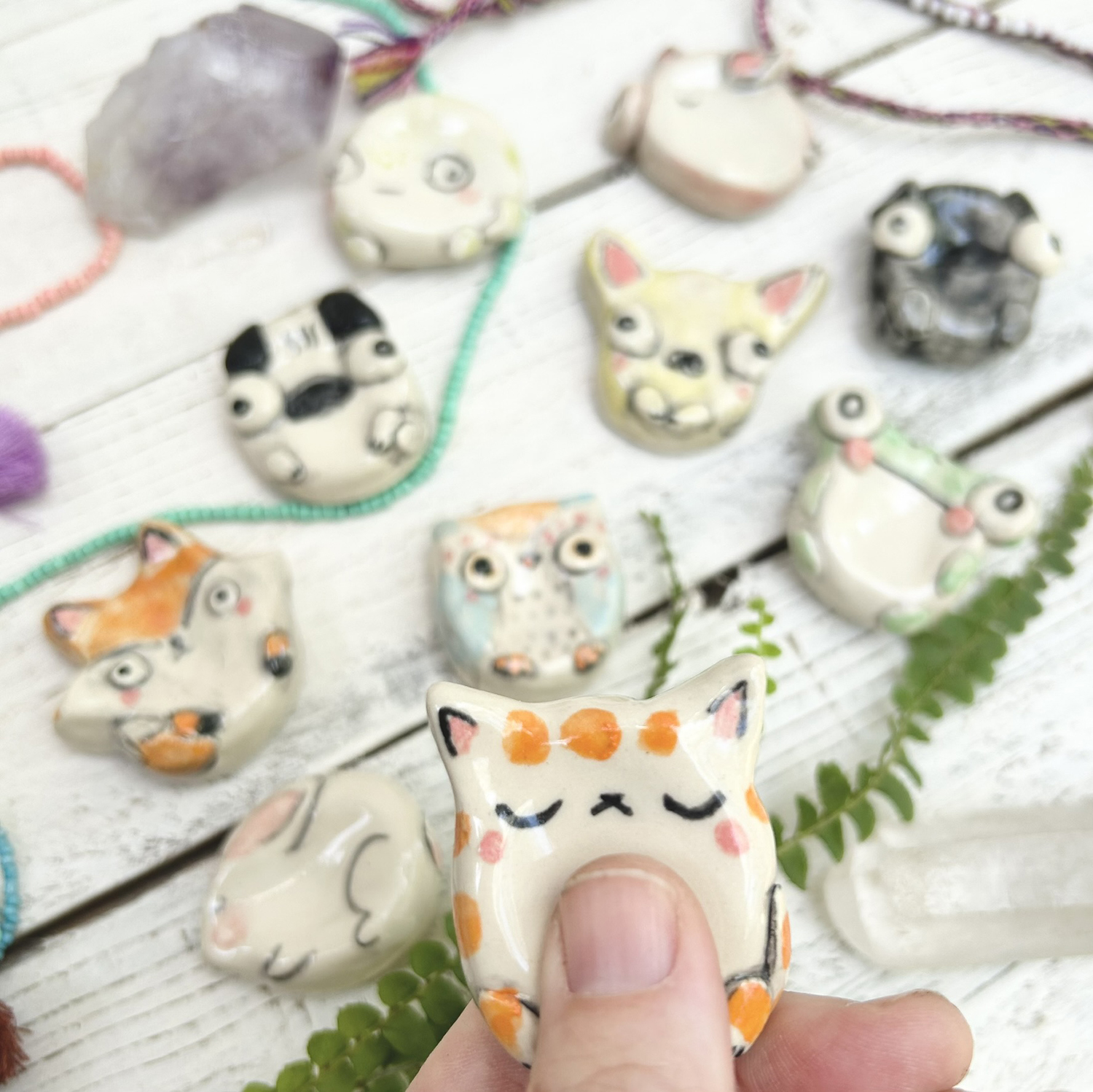 Hand holding a small ceramic cat figurine with orange spots, surrounded by other similar figurines on a white wooden surface with green leaves