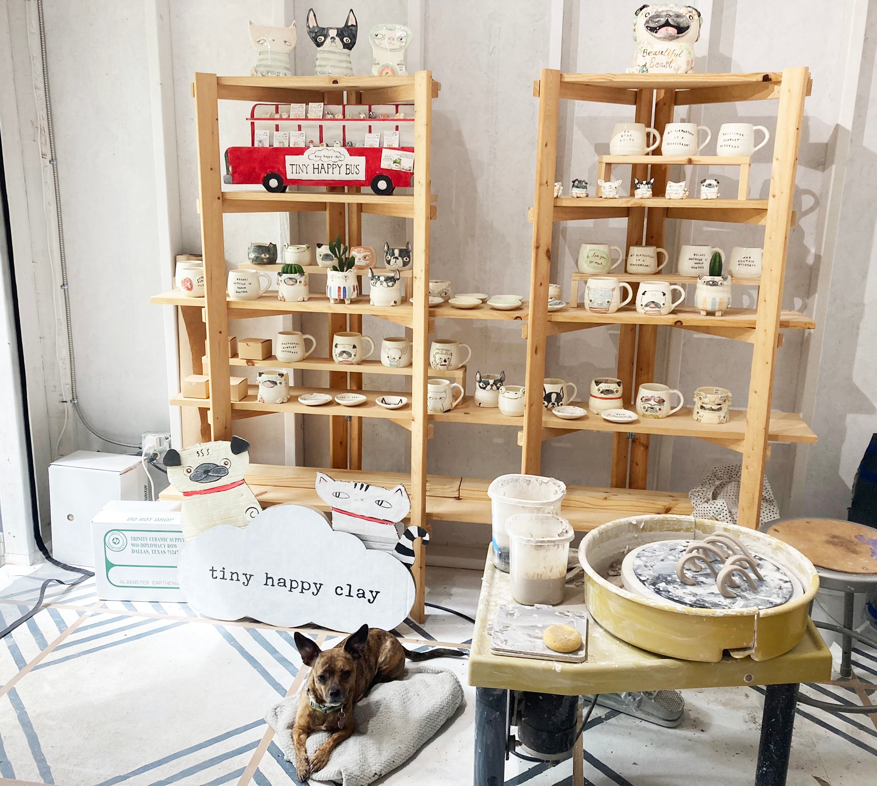 Display shelves with pottery, a dog lying on a cushion, and a sign reading 'tiny happy clay' in a room.