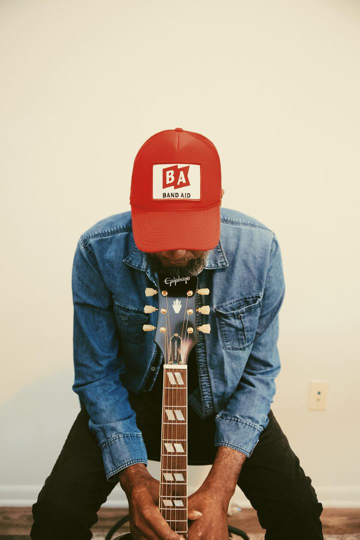 Person wearing a red cap, denim jacket, and playing an electric guitar against a plain wall.
