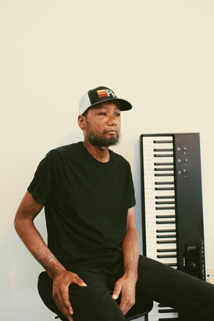 Man with beard and cap sitting next to a keyboard, wearing a black t-shirt, against a plain light background.