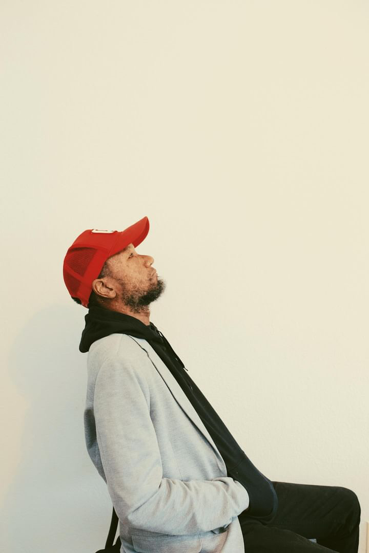 Man wearing a red cap, gray jacket, and black scarf, sitting with eyes closed against a plain light background.