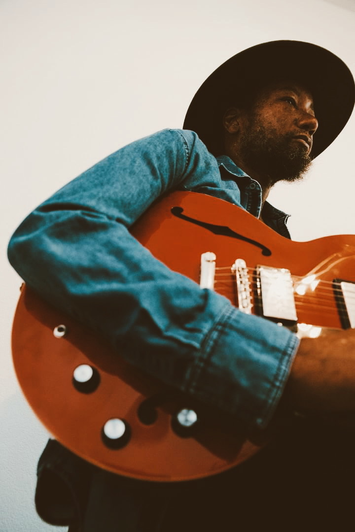 Person wearing a hat and denim shirt holding an orange electric guitar, looking to the side.