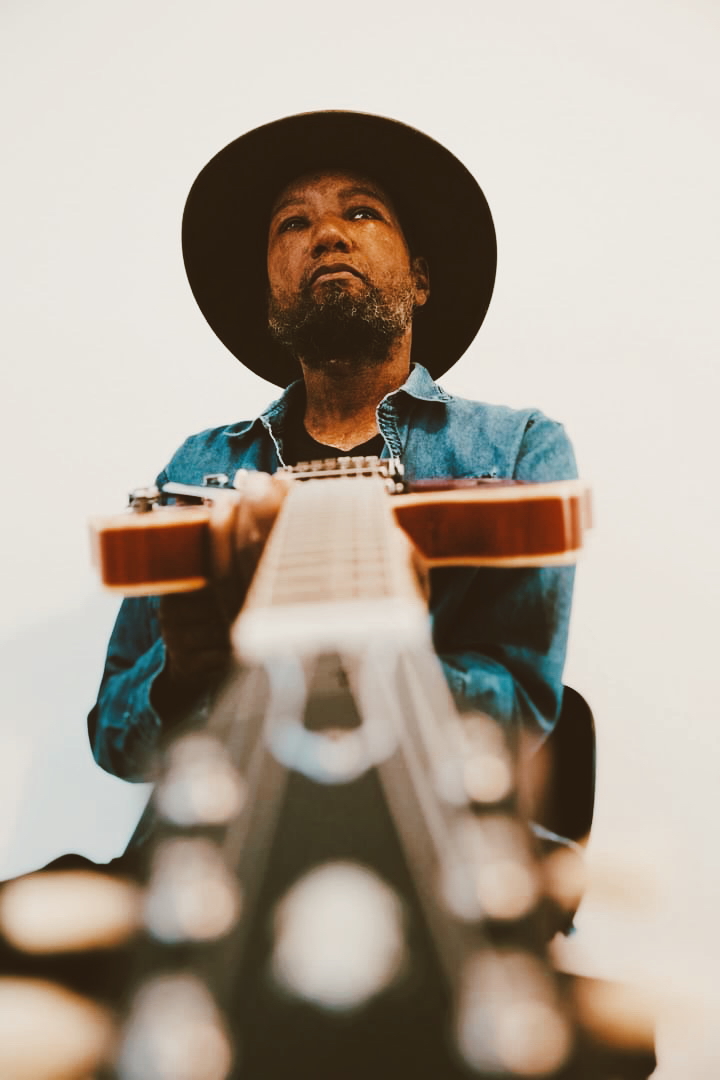 Person wearing a hat and denim jacket looking down at a guitar, with a plain background.