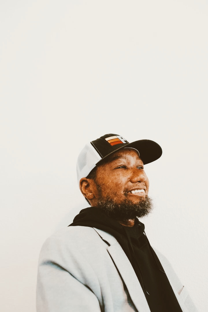 Man wearing a cap and gray blazer smiling, facing right, against a plain white background.