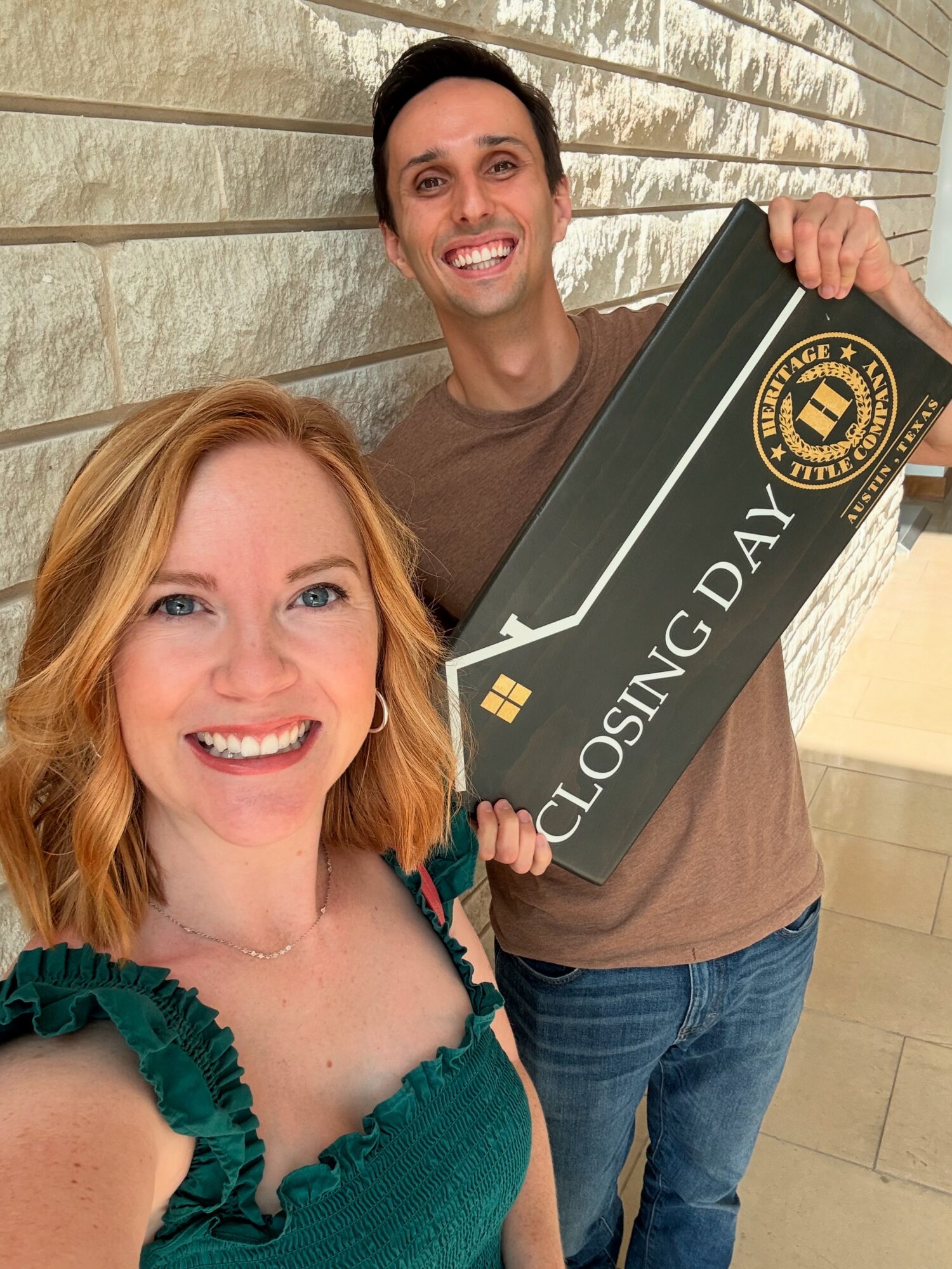 Two smiling people holding a large sign that says 'Closing Day' in front of a textured wall.