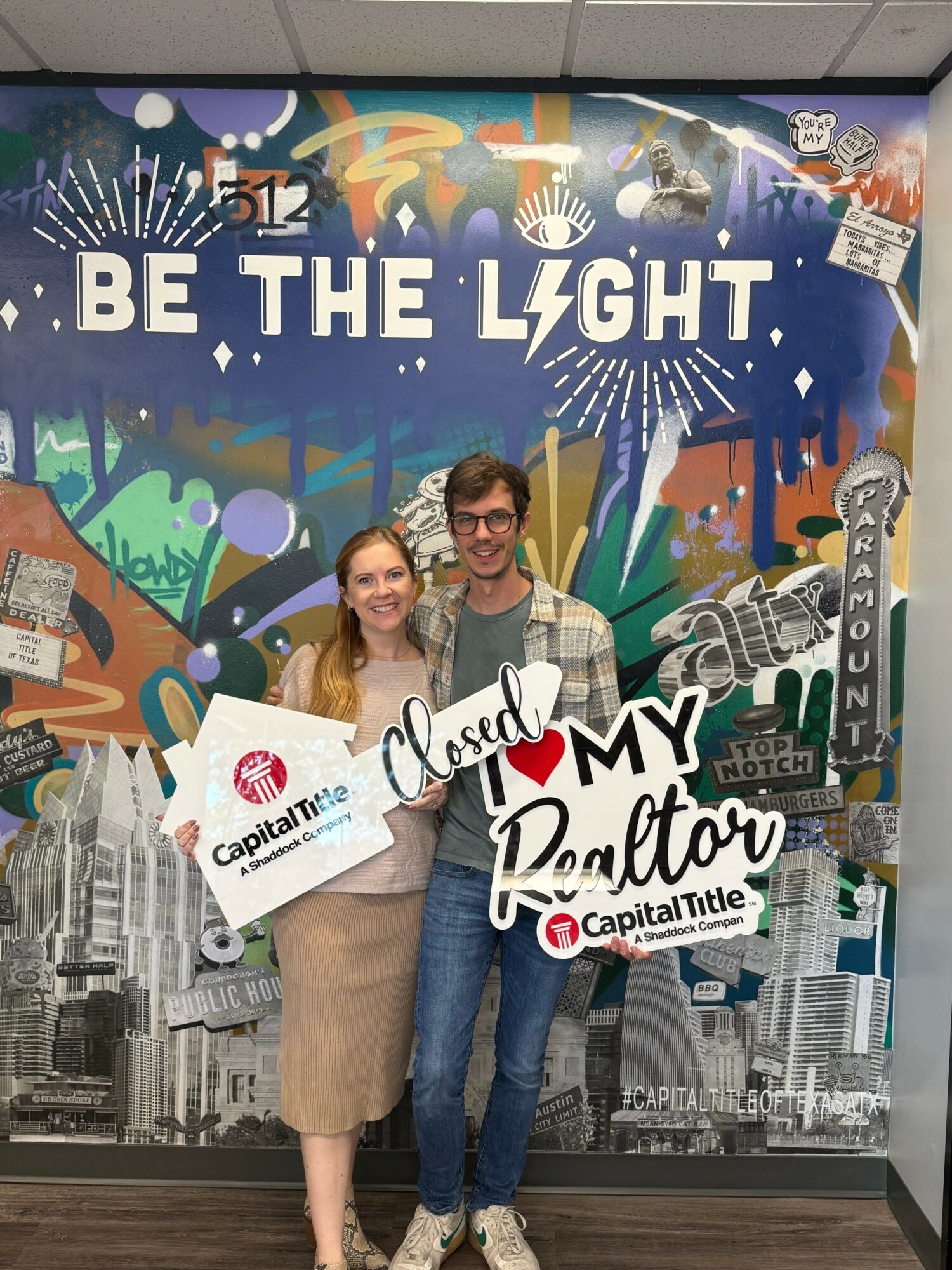 Two people holding signs in front of a colorful mural with the text 'BE THE LIGHT'.