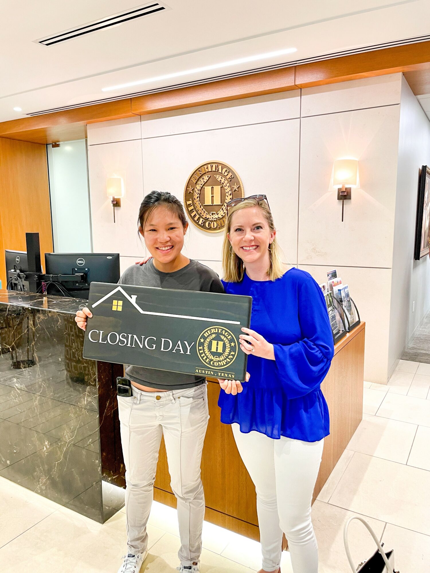 Two women smiling at a reception desk holding a sign that says 'Closing Day' in a professional office setting.