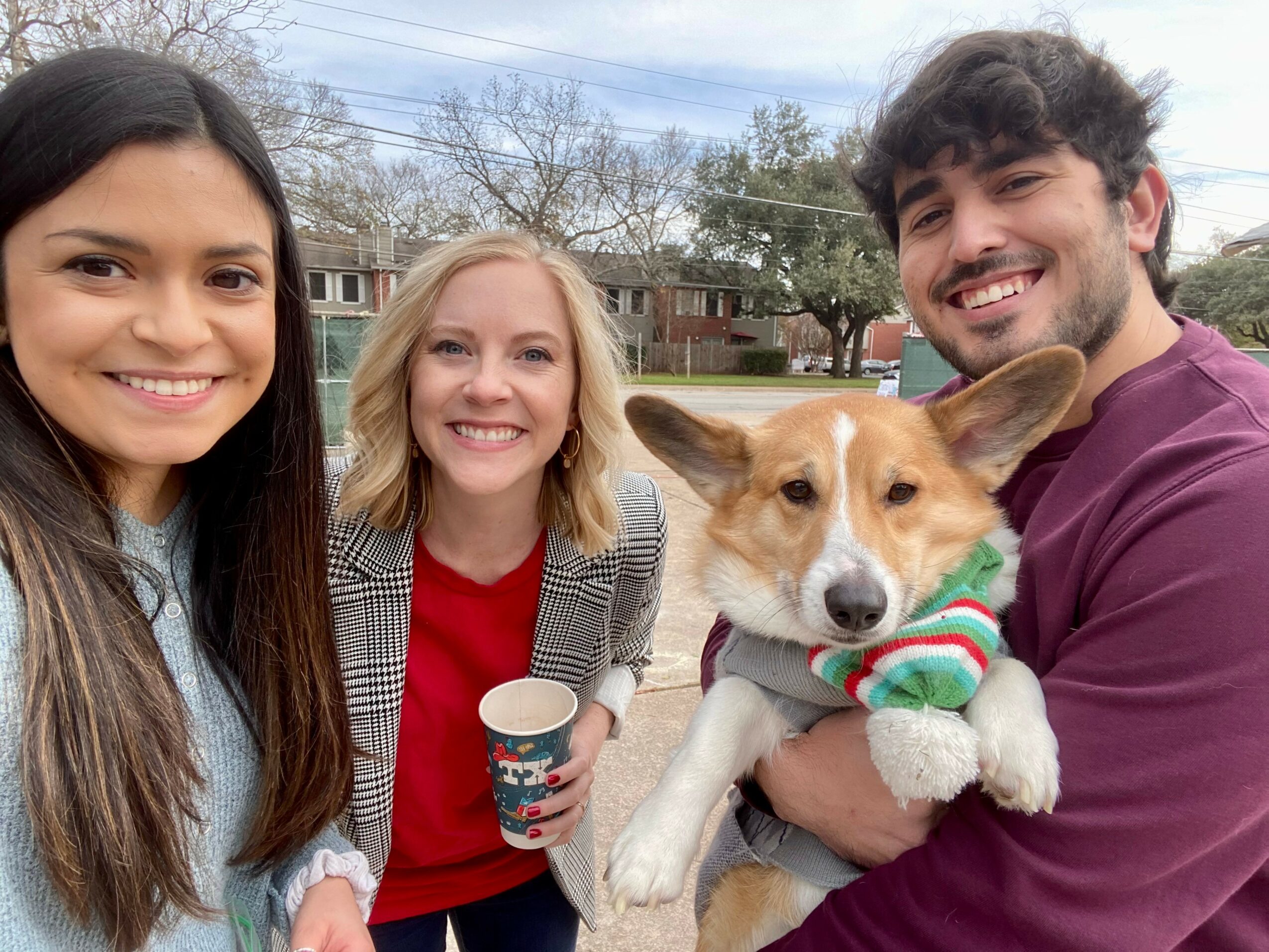 Three people and a dog outdoors, smiling, with houses and trees in background.