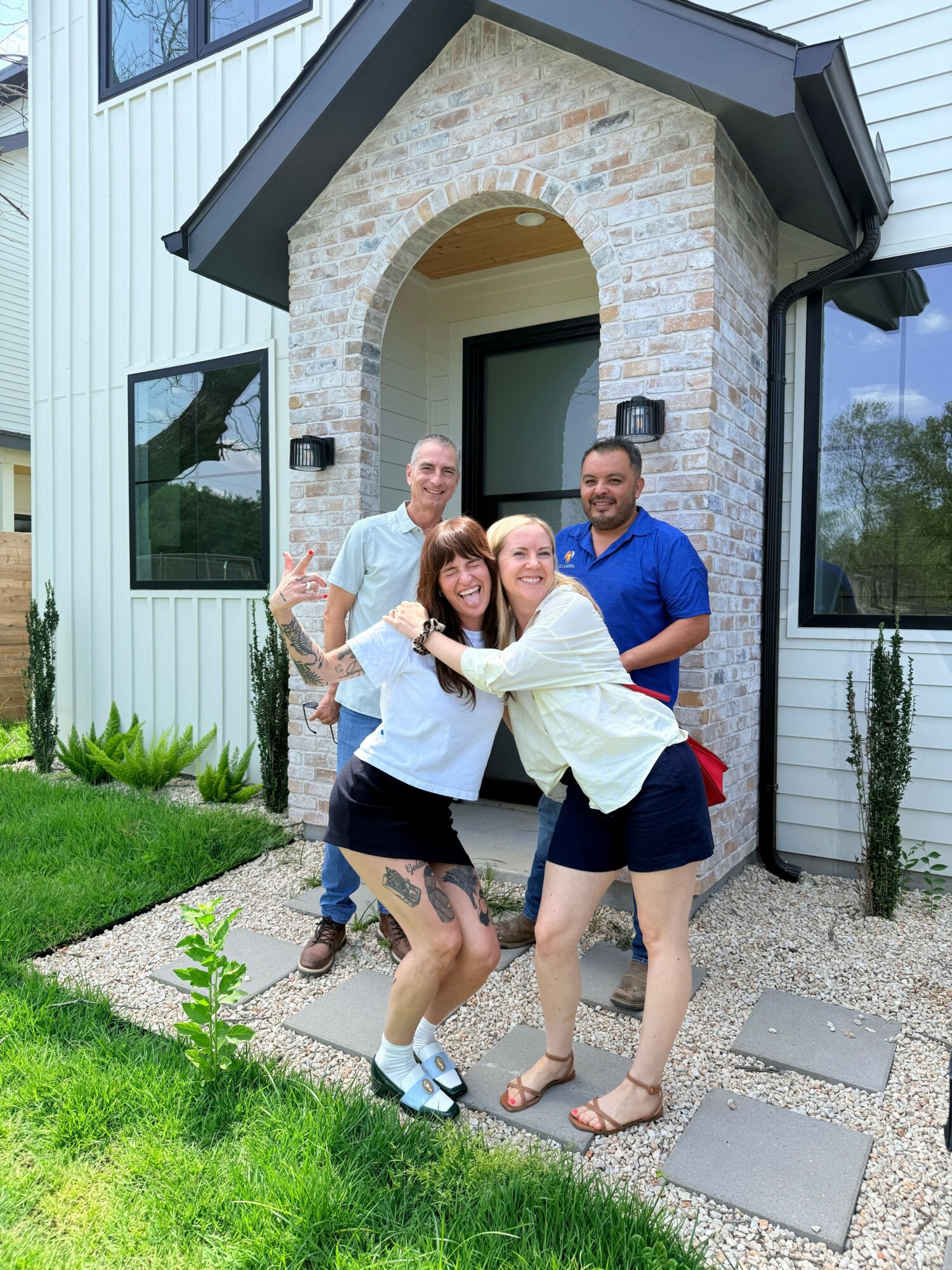 Four people standing outside a house, smiling and posing for a photo, with a stone archway and window behind them.