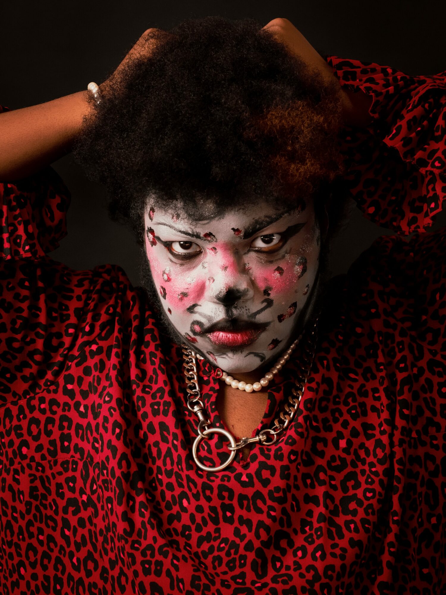 Person with face makeup, wearing a red and black leopard print outfit, jewelry, and a serious expression.
