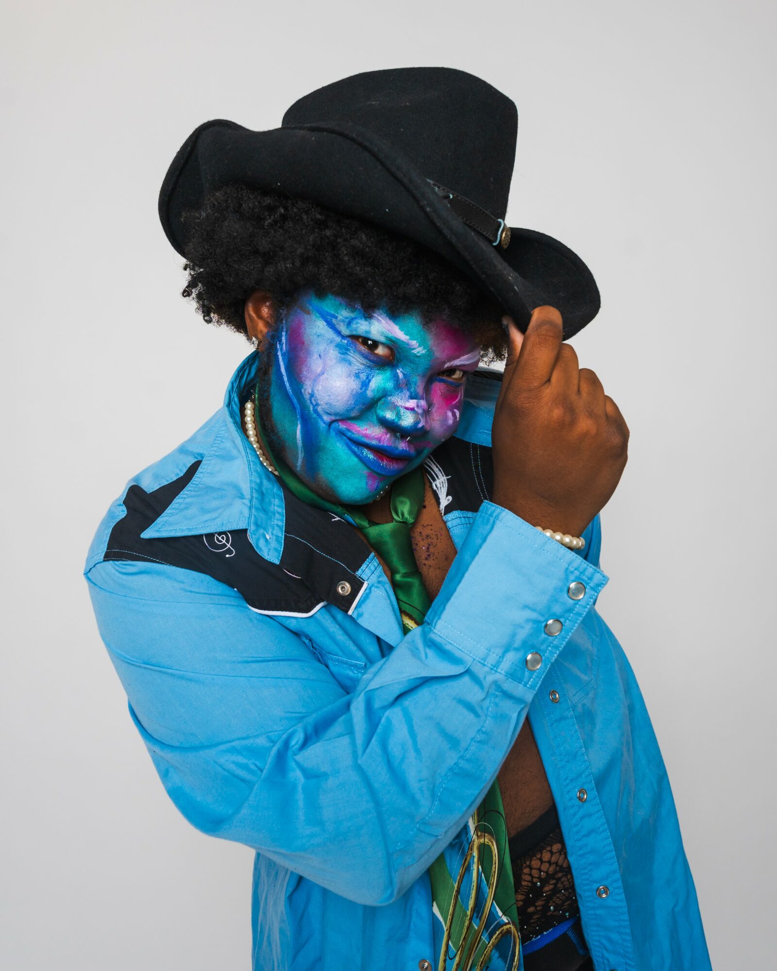 Person with blue face paint, wearing a black hat and blue jacket, holding the hat's brim, against a plain background.