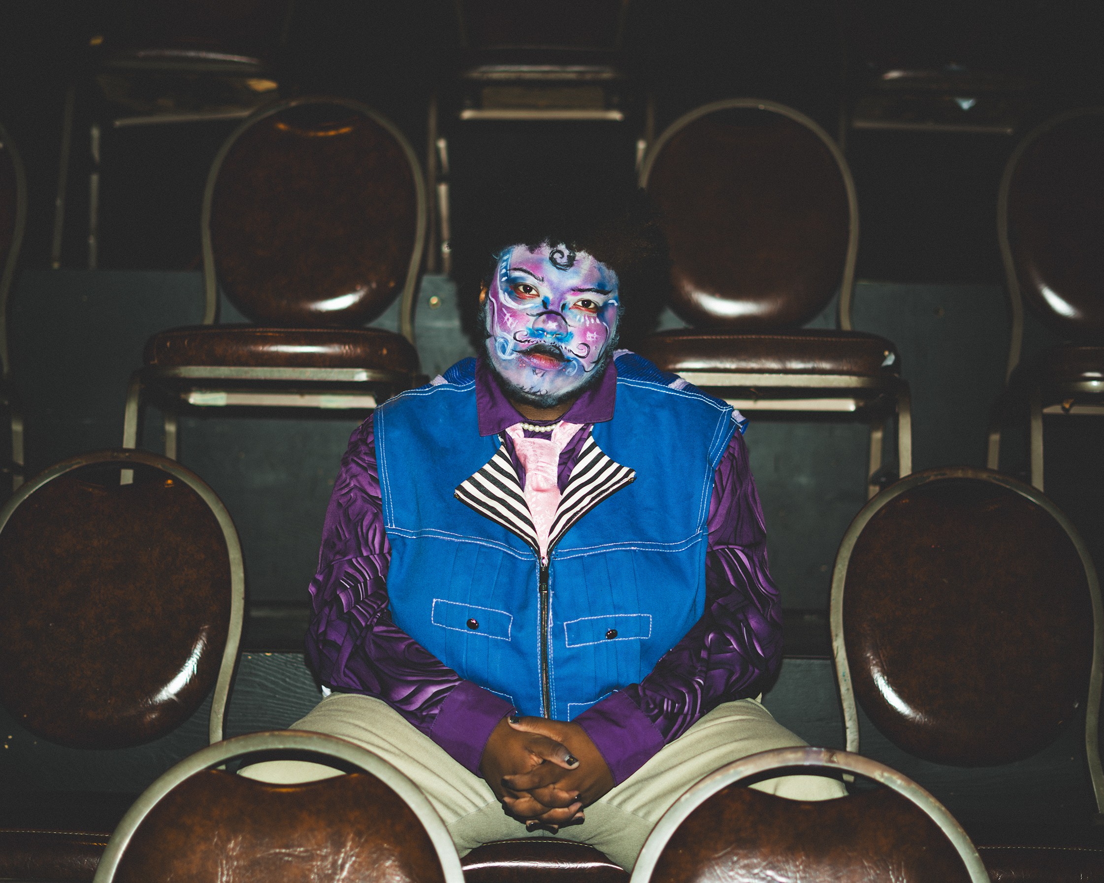 Person with clown makeup sitting alone in a theater with empty chairs around them.