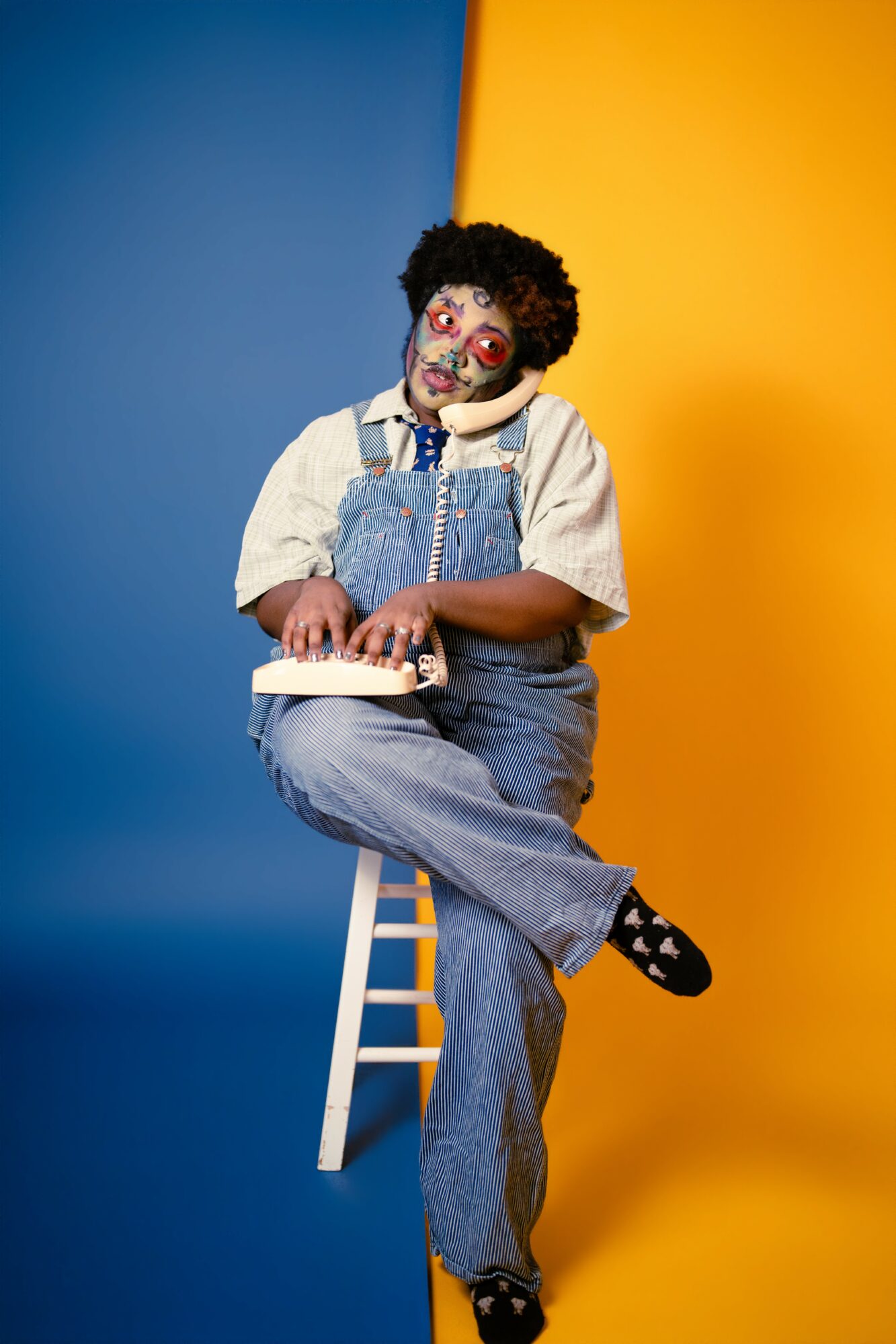 Clown with curly hair, painted face, and clown nose, sitting on a stool with crossed legs, holding a book, against colorful background.