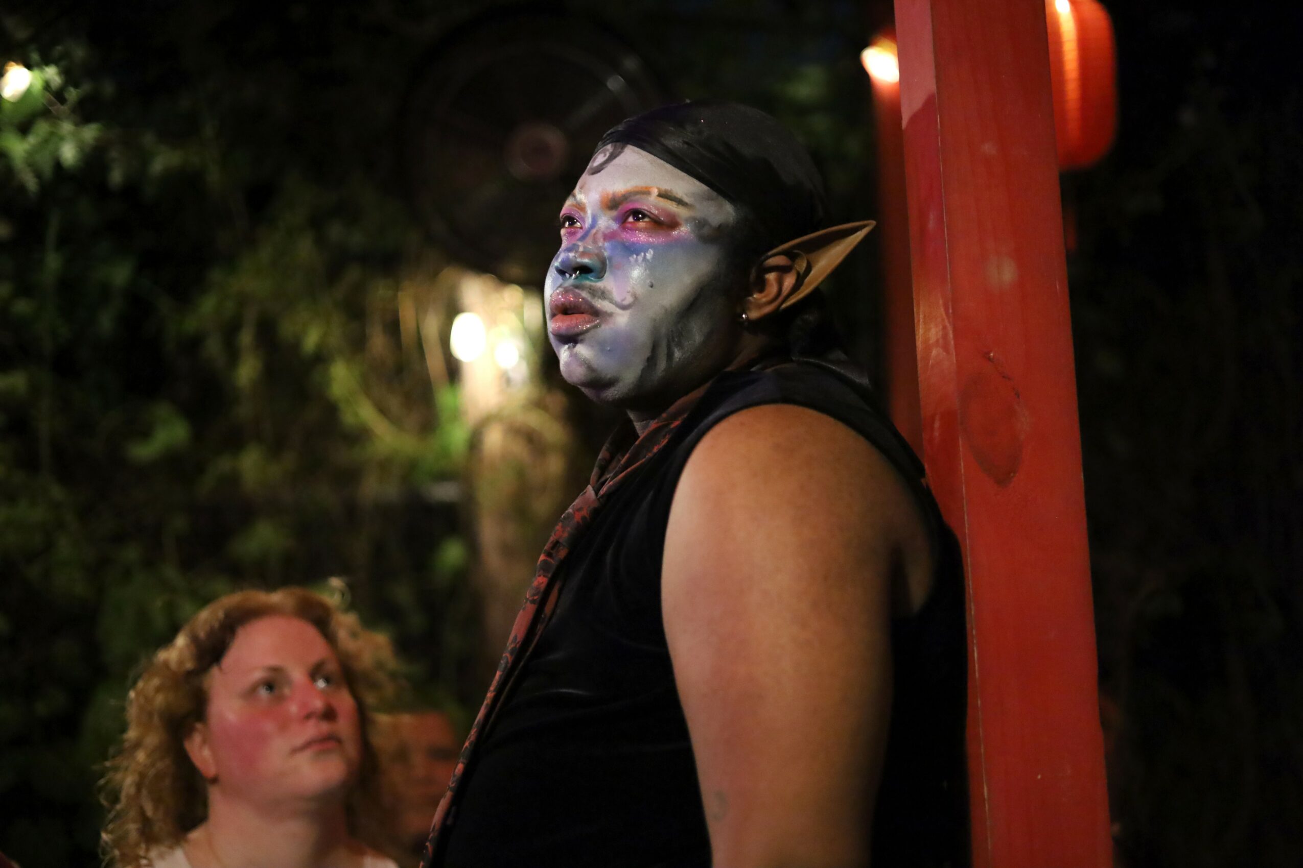 Person with elaborate face makeup and elf-like ears leaning against a red post outdoors at night.