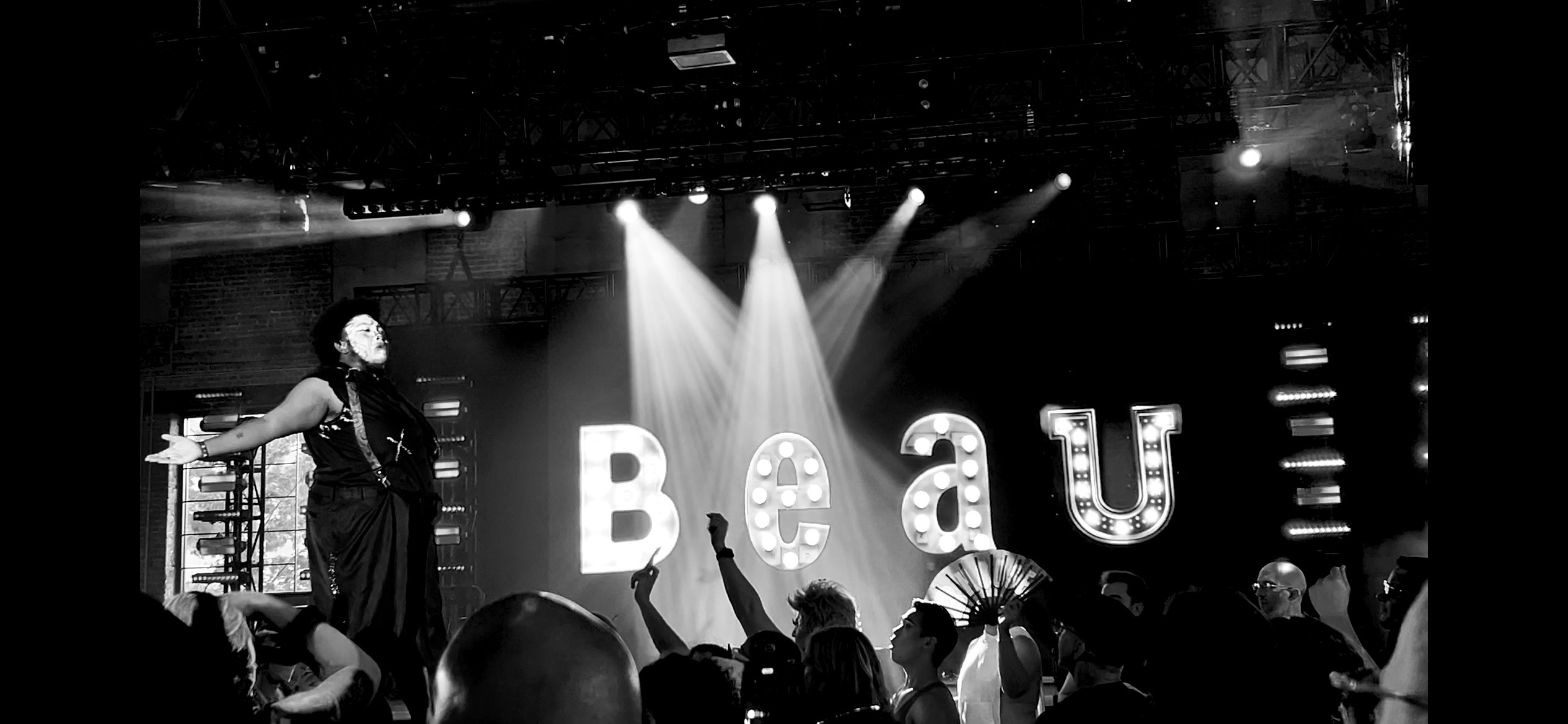 Stage with large illuminated letters spelling 'Beau' and a performer on the left, audience in foreground.