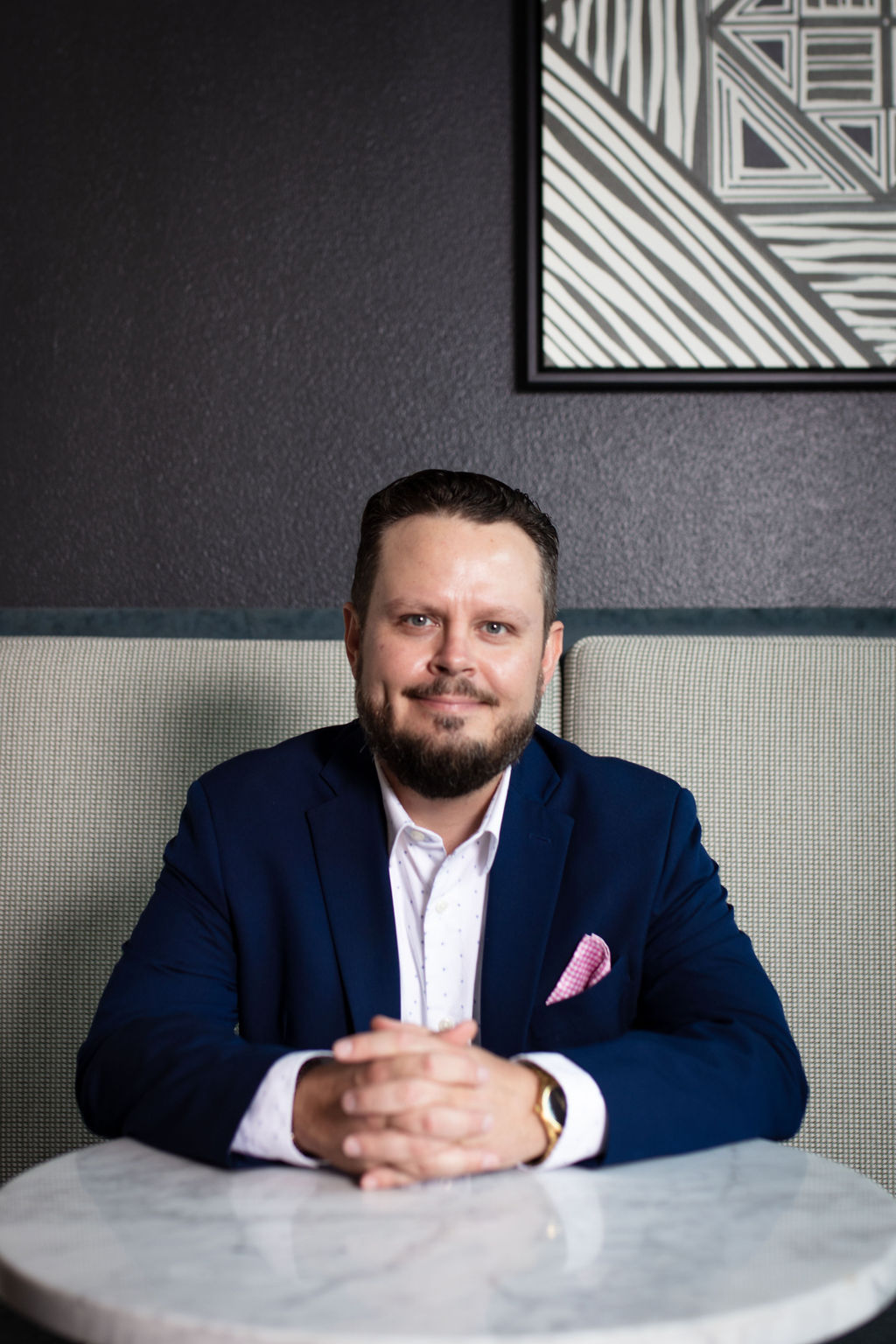 Man with dark hair and beard in a suit sitting at a table, smiling, with a dark wall and framed artwork behind him.