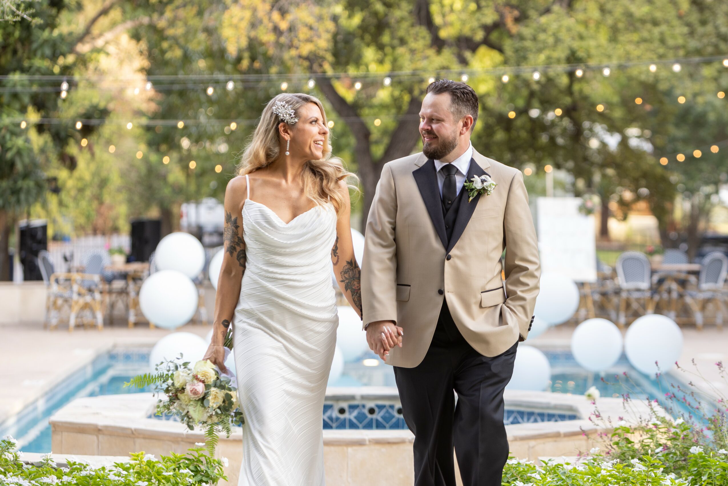 Bride and groom holding hands, smiling, outdoors near a pool with string lights and trees in the background.