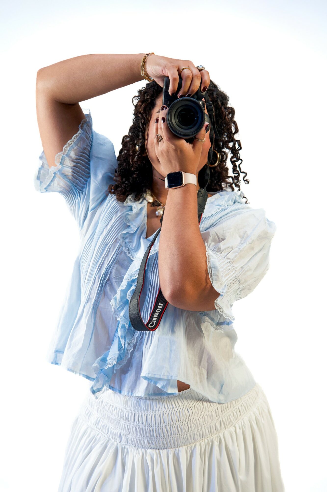 Person with curly hair holding a camera up to their face, wearing a light blue and white striped shirt and white skirt.