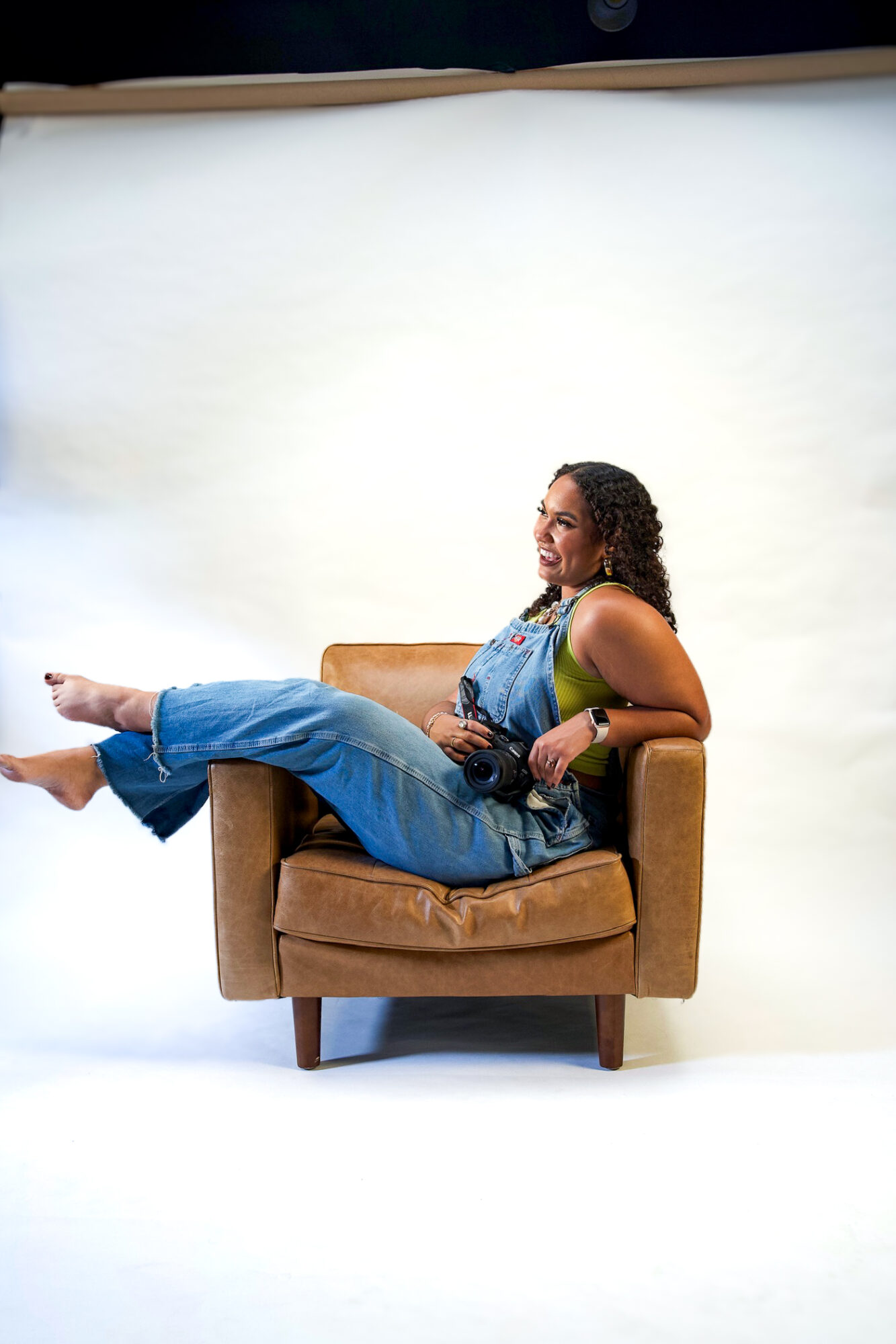 Person sitting on a brown armchair with legs extended, smiling, against a plain white background.