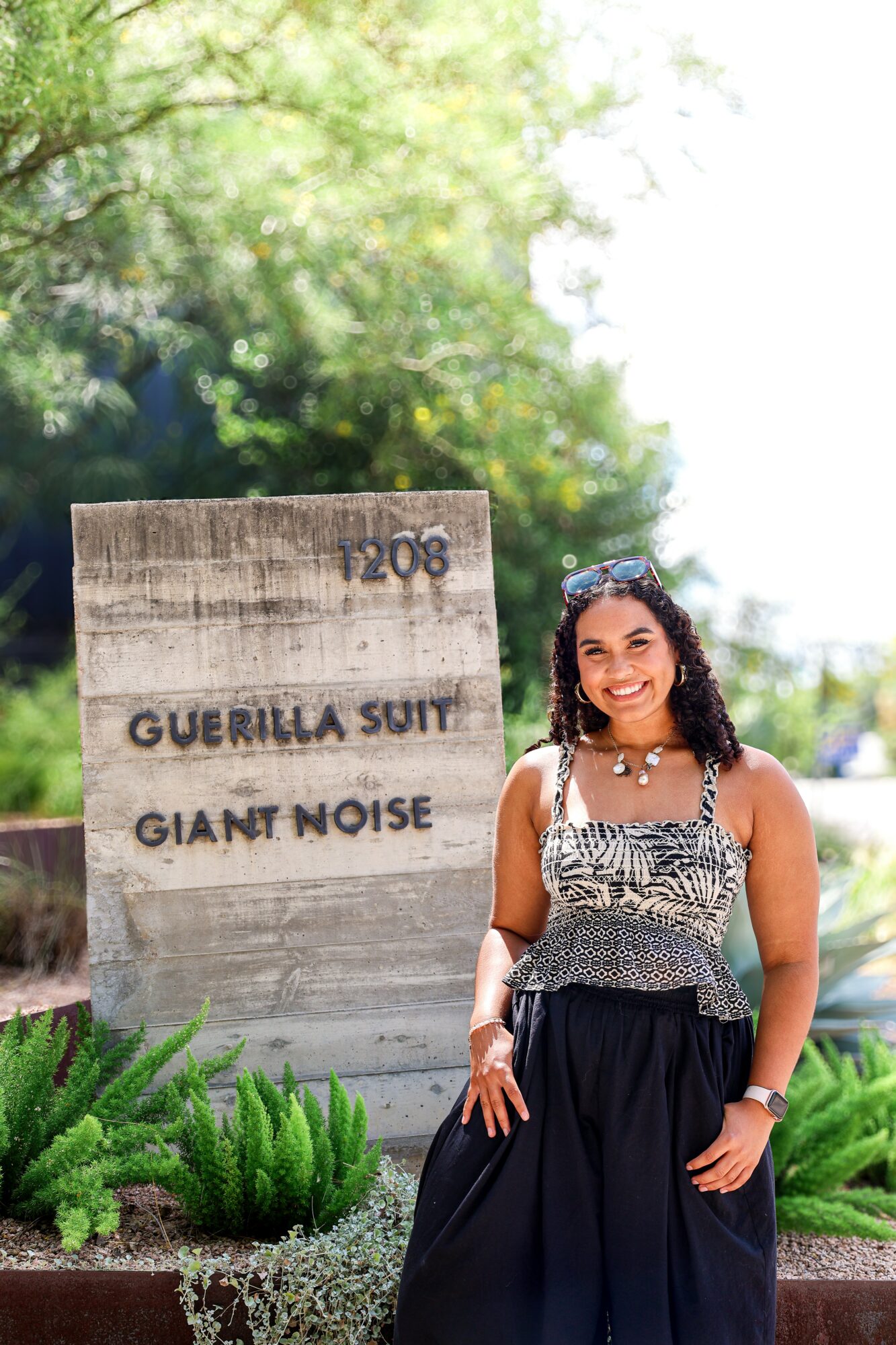 Smiling woman standing outdoors near a wooden sign with text, surrounded by greenery and trees.