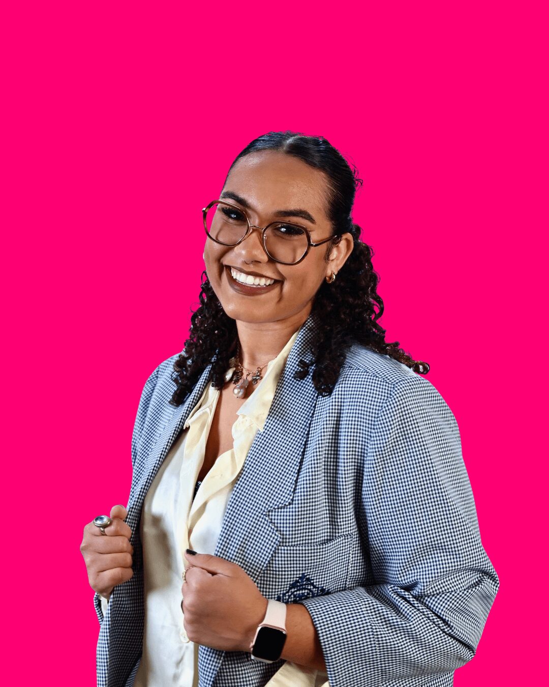 Woman with glasses smiling, wearing a blazer and a watch, against a bright pink background.
