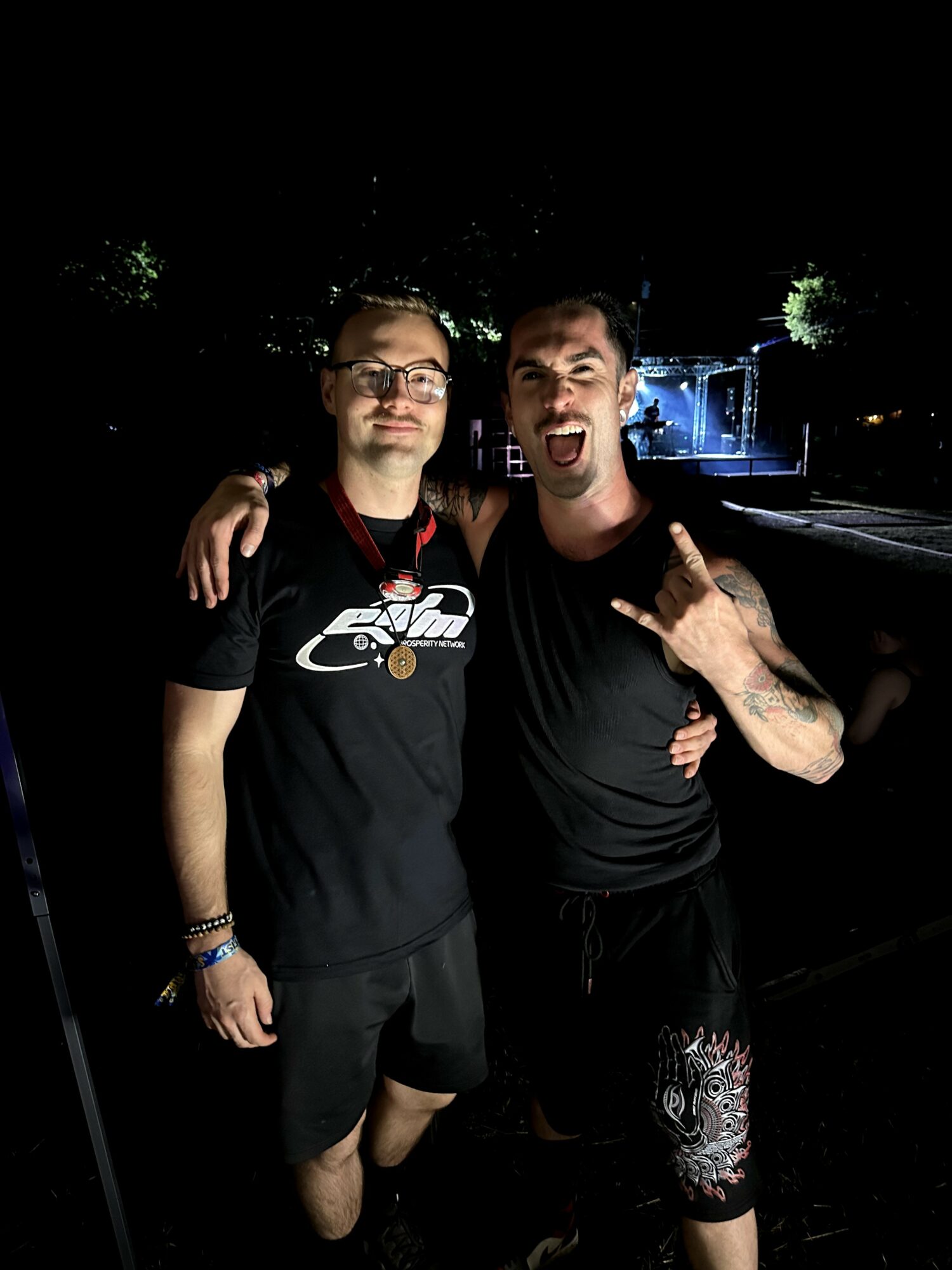Two men standing outdoors at night, one with arm around the other, smiling and making a gesture with his hand.