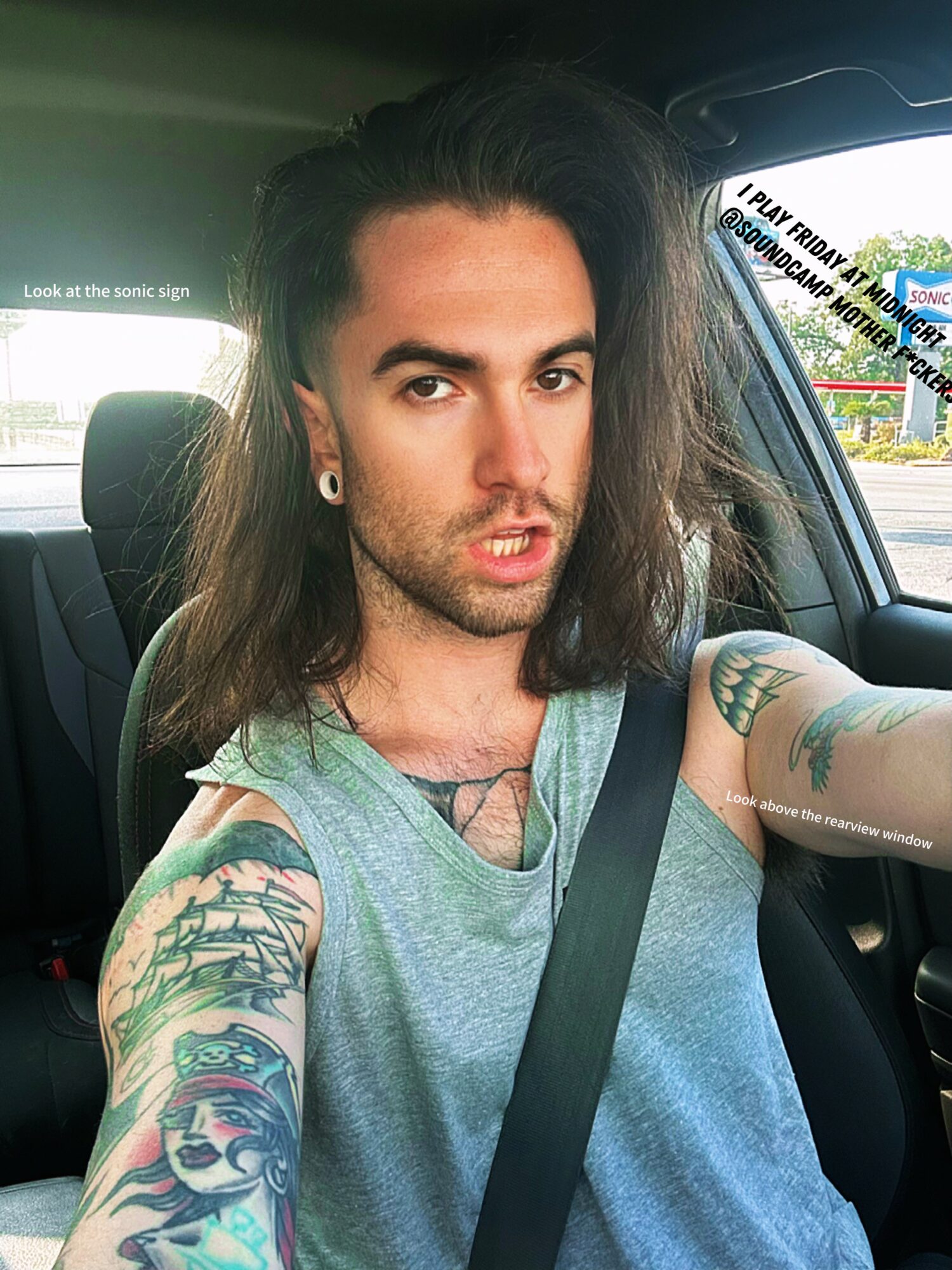 Person with long hair and tattoos on arms taking a selfie inside a car, wearing a gray tank top.