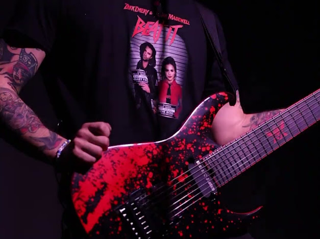 Person holding a red and black electric guitar, wearing a black T-shirt with a graphic of two women and text, tattooed arm visible.