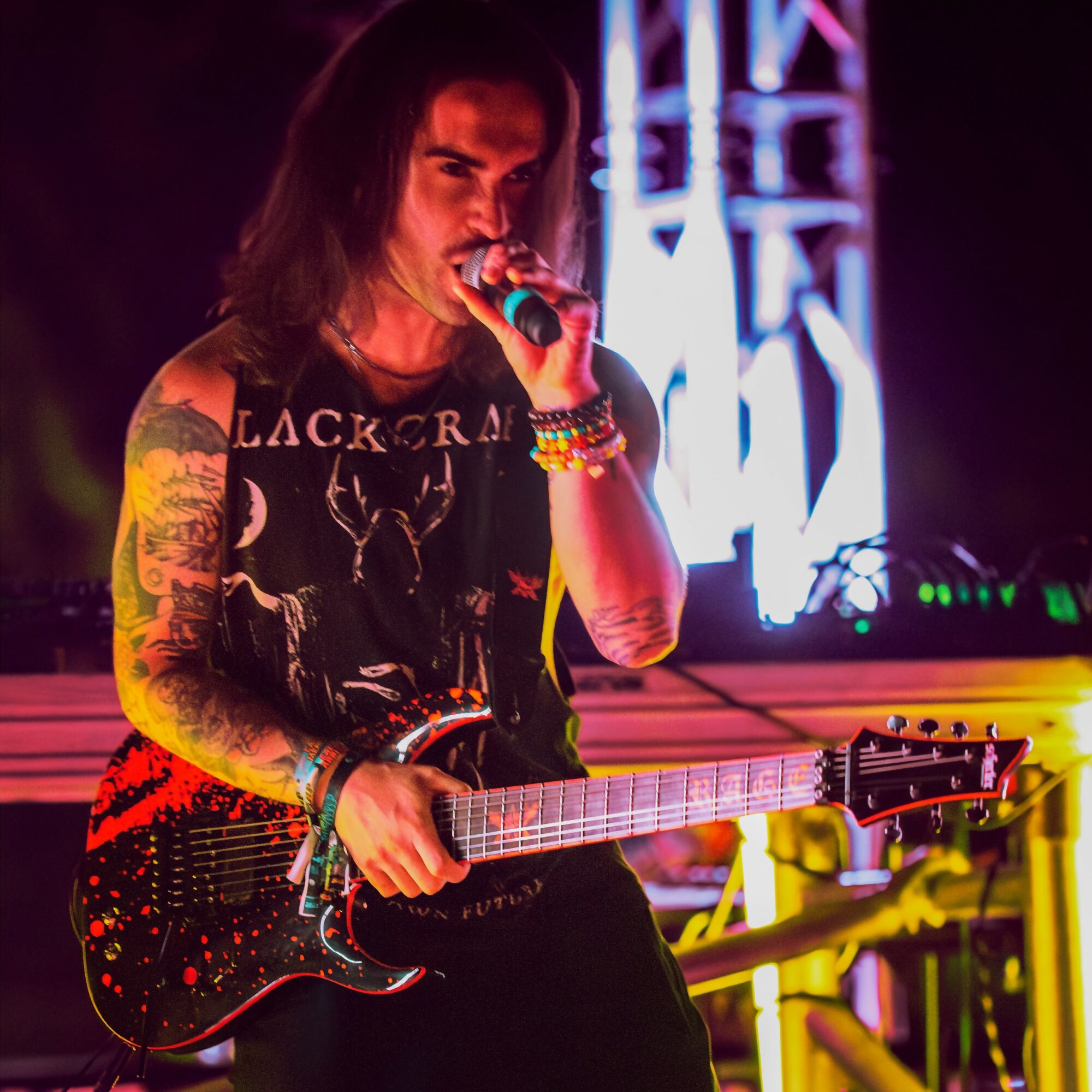 Female musician with tattoos, holding a guitar and singing into a microphone on stage with colorful lighting.