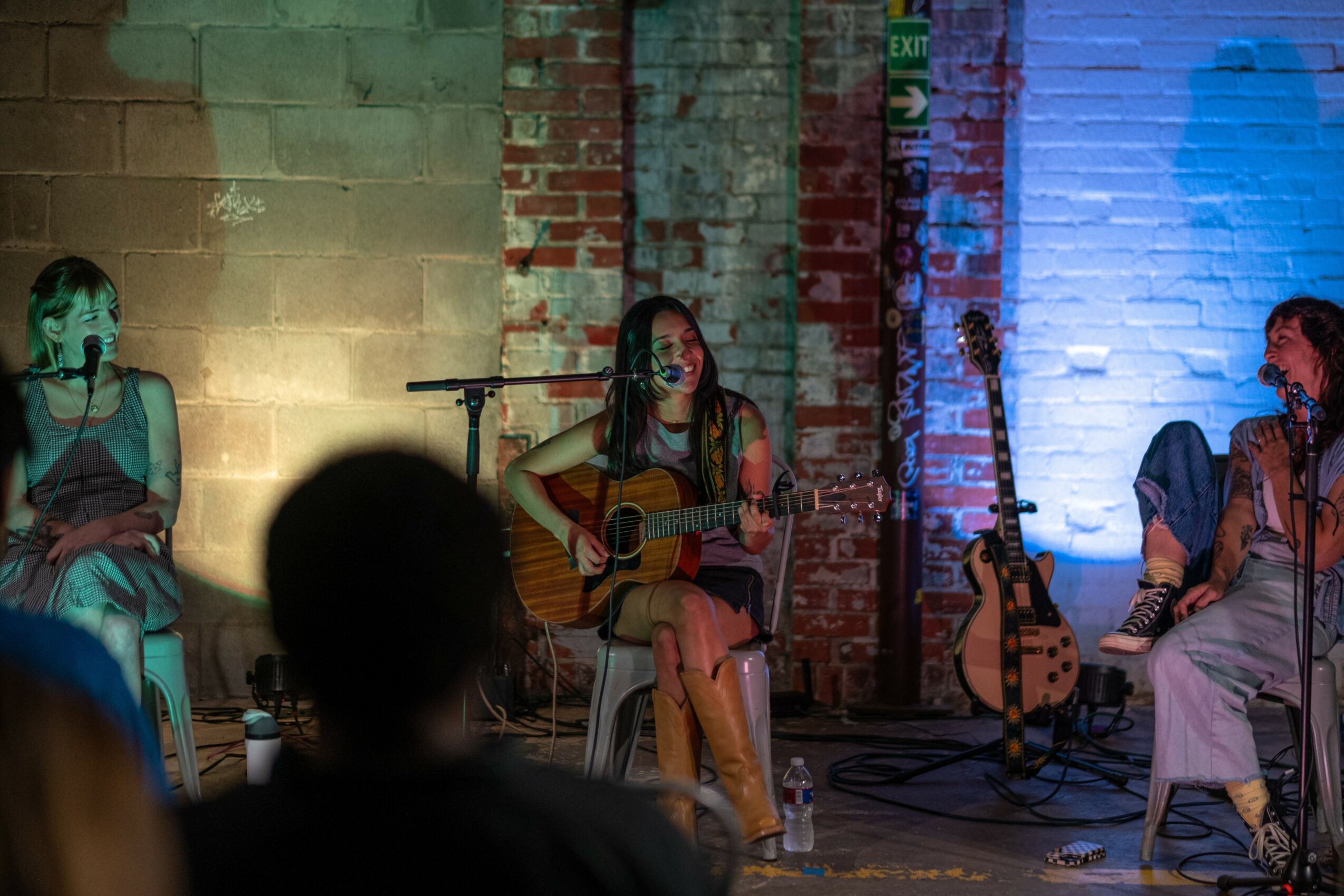 Three women perform on stage with guitars, seated against a brick wall, with audience silhouettes in foreground.