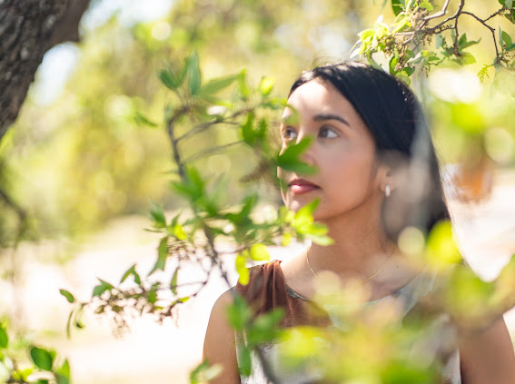 Young woman with dark hair outdoors among green leaves, looking to the side, sunlight filtering through trees.