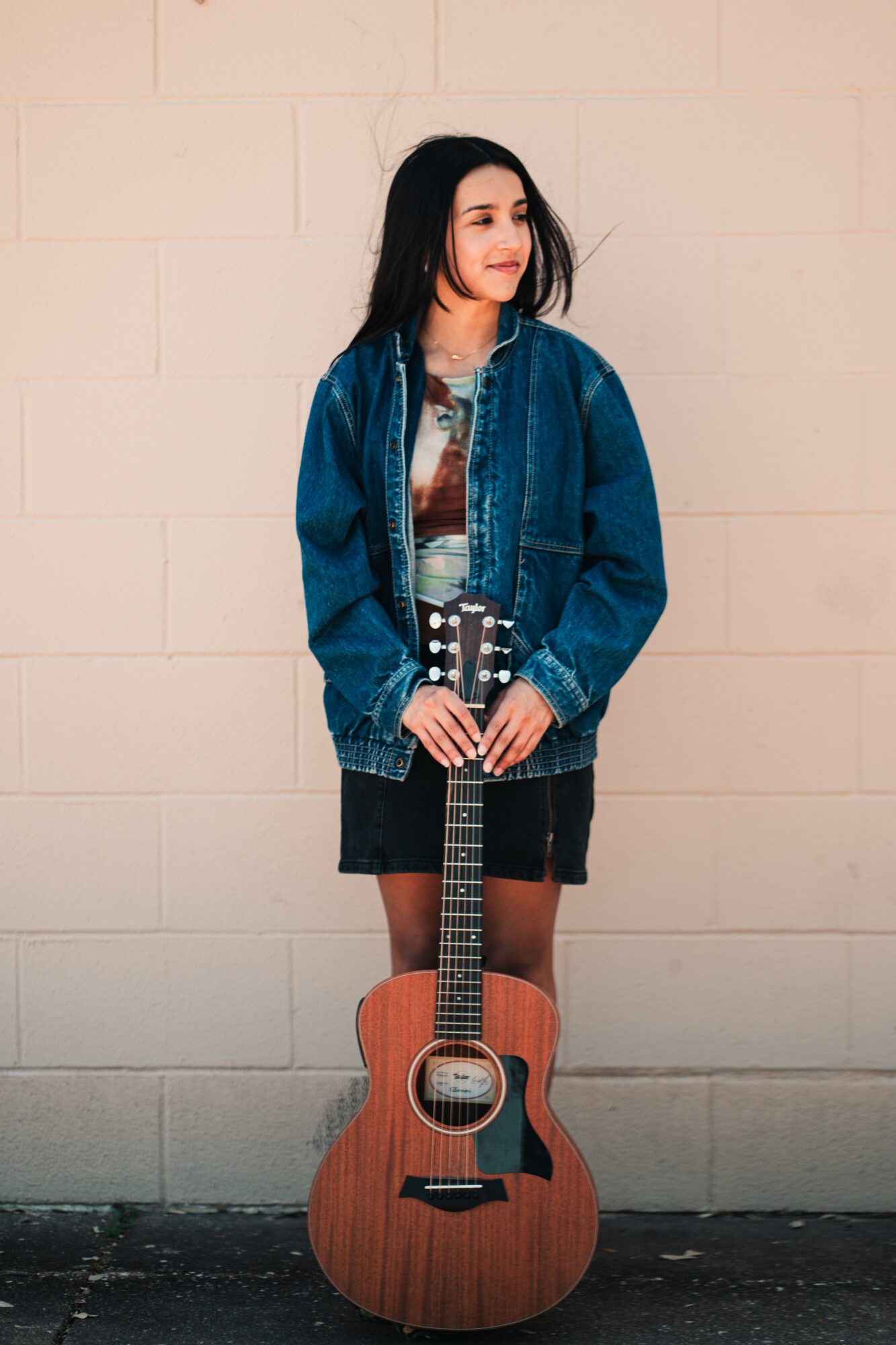 Young woman with long dark hair in a denim jacket holding an acoustic guitar, standing against a plain wall.