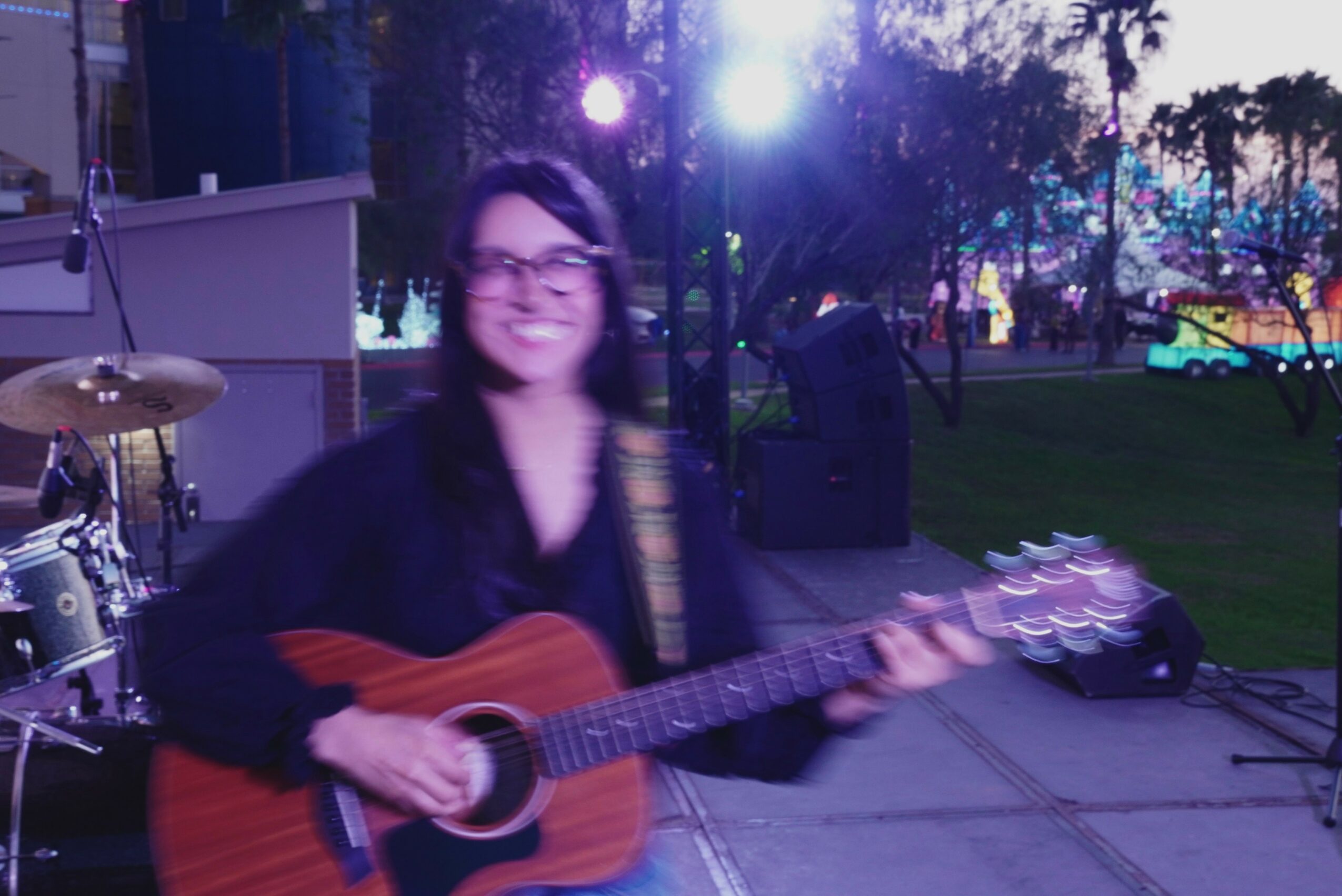 Person playing guitar outdoors at night with colorful lights and trees in background.