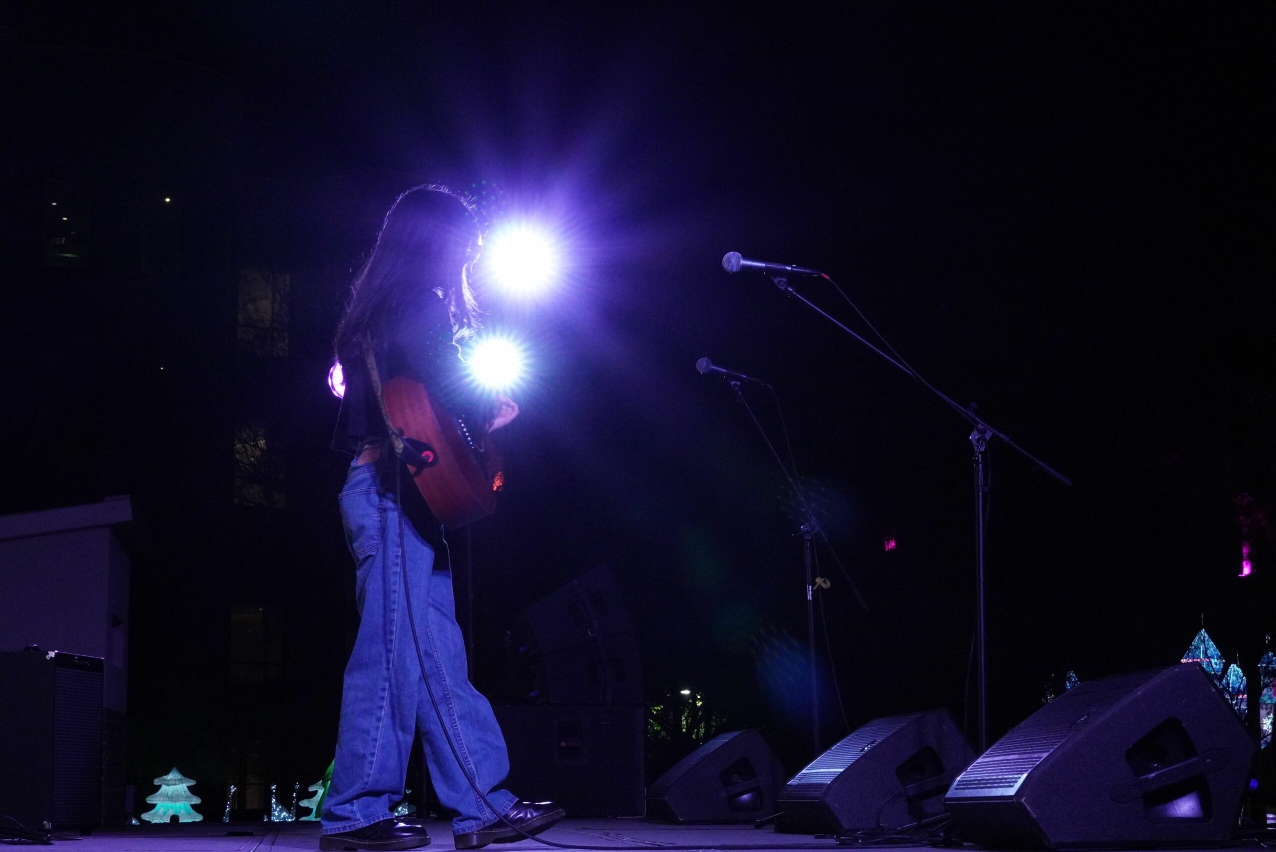 Person playing guitar on stage with bright lights and microphones, dark background, stage equipment visible.