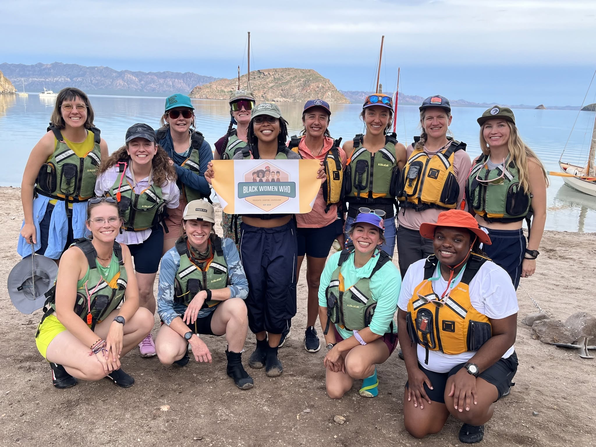 Group of twelve people in life jackets and hats on a beach with boats and water in background.