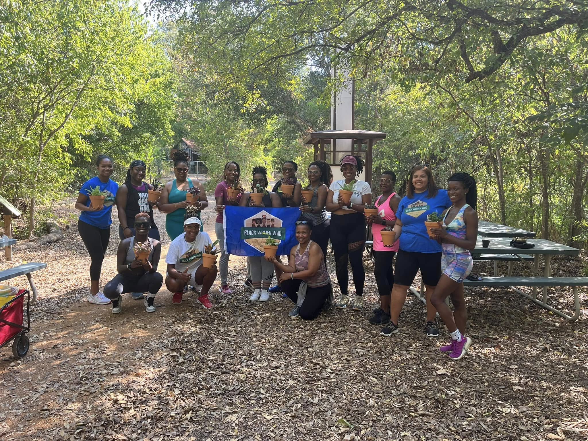Group of people outdoors in a wooded area, some holding a sign, posing for a photo, with trees and picnic tables in background.