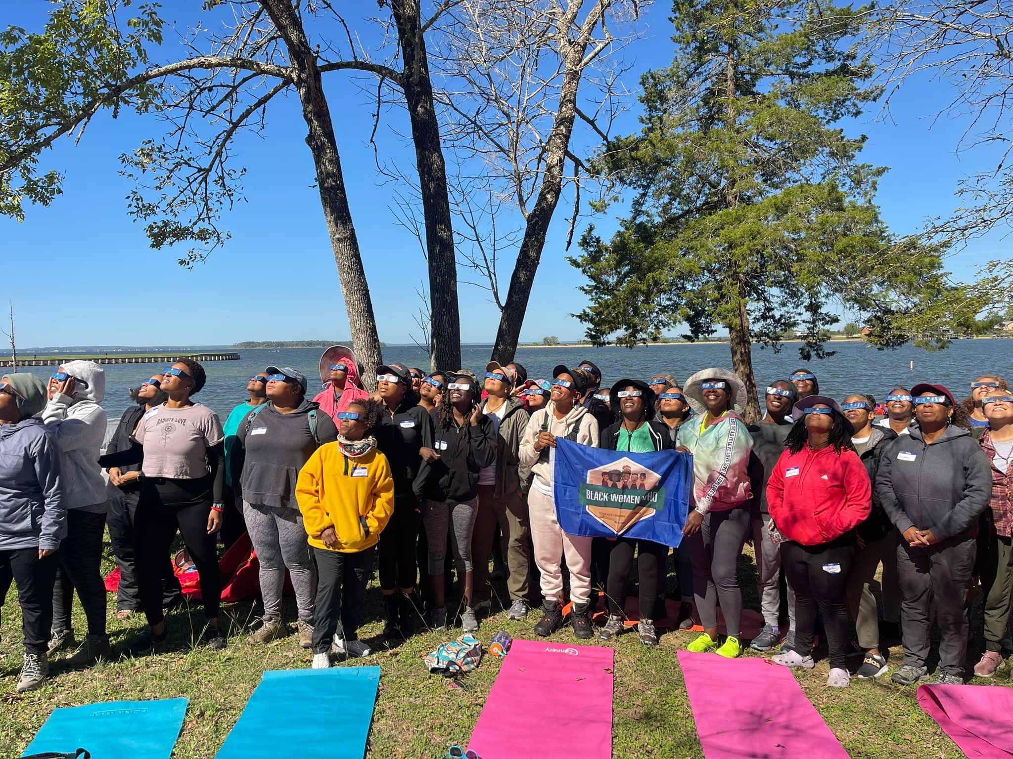 Group of people outdoors near water with trees, some holding a blue flag, on a sunny day.