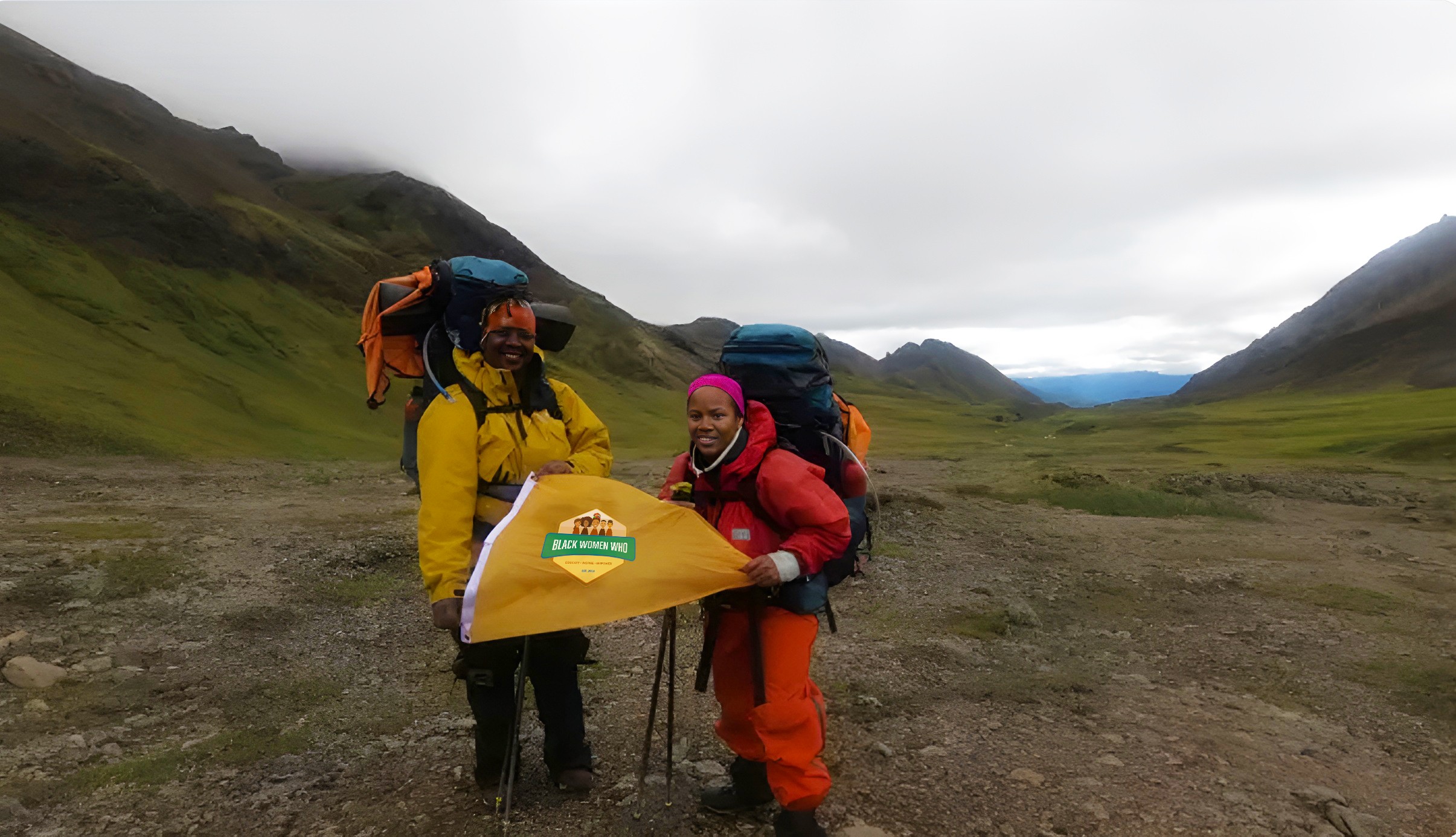 Two hikers with backpacks standing in a valley with mountains in the background, holding a yellow flag or banner.