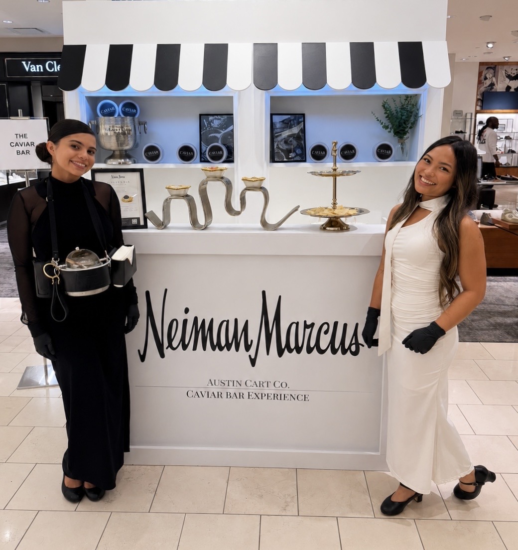 Two women stand on either side of a Neiman Marcus display with a white and black striped canopy, smiling. One wears black, the other white, both with gloves.