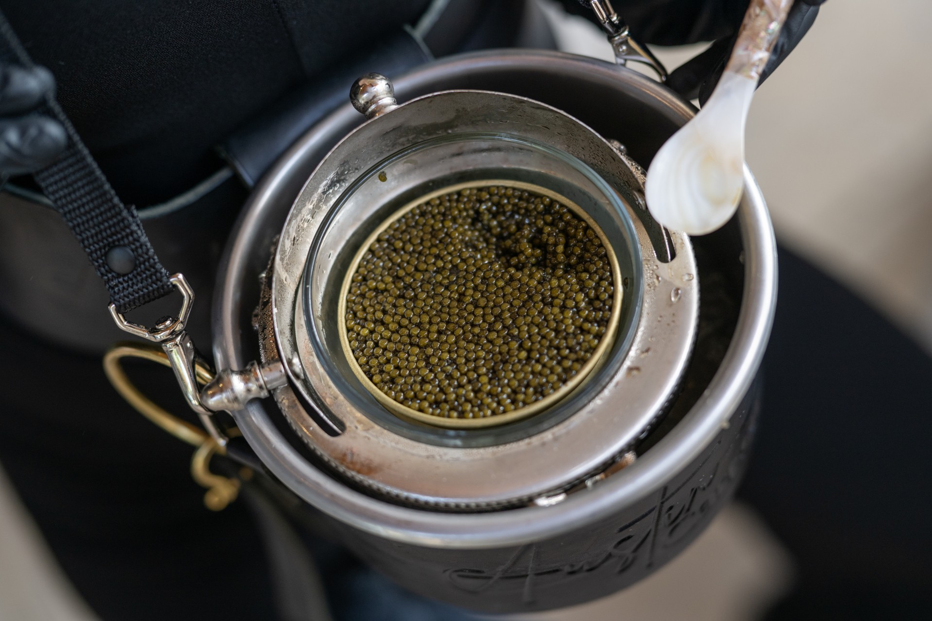 A glass cup with green and black peppercorns inside, placed on a metal container with a white spoon nearby.