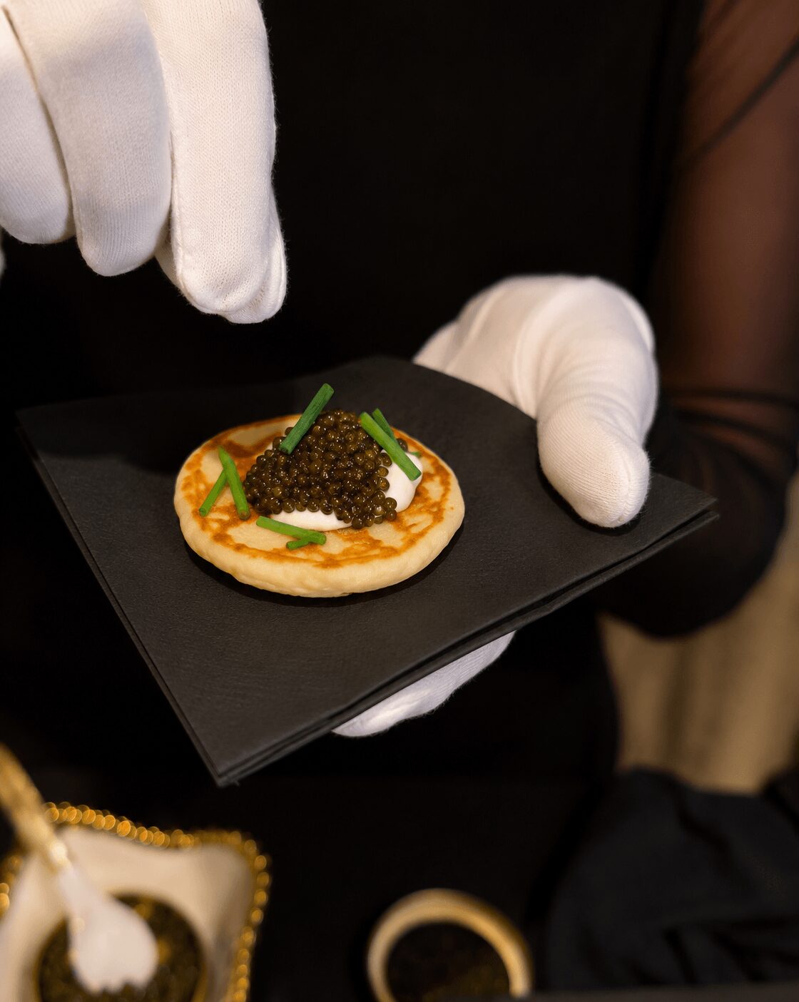 Person wearing white gloves holding a small dish with caviar, garnished with green herbs on a black tray.