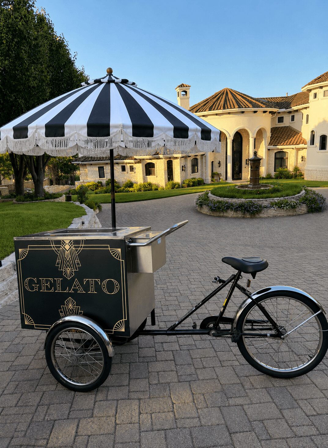 Gelato cart attached to a bicycle with a striped umbrella, set in a courtyard with buildings and greenery in the background.