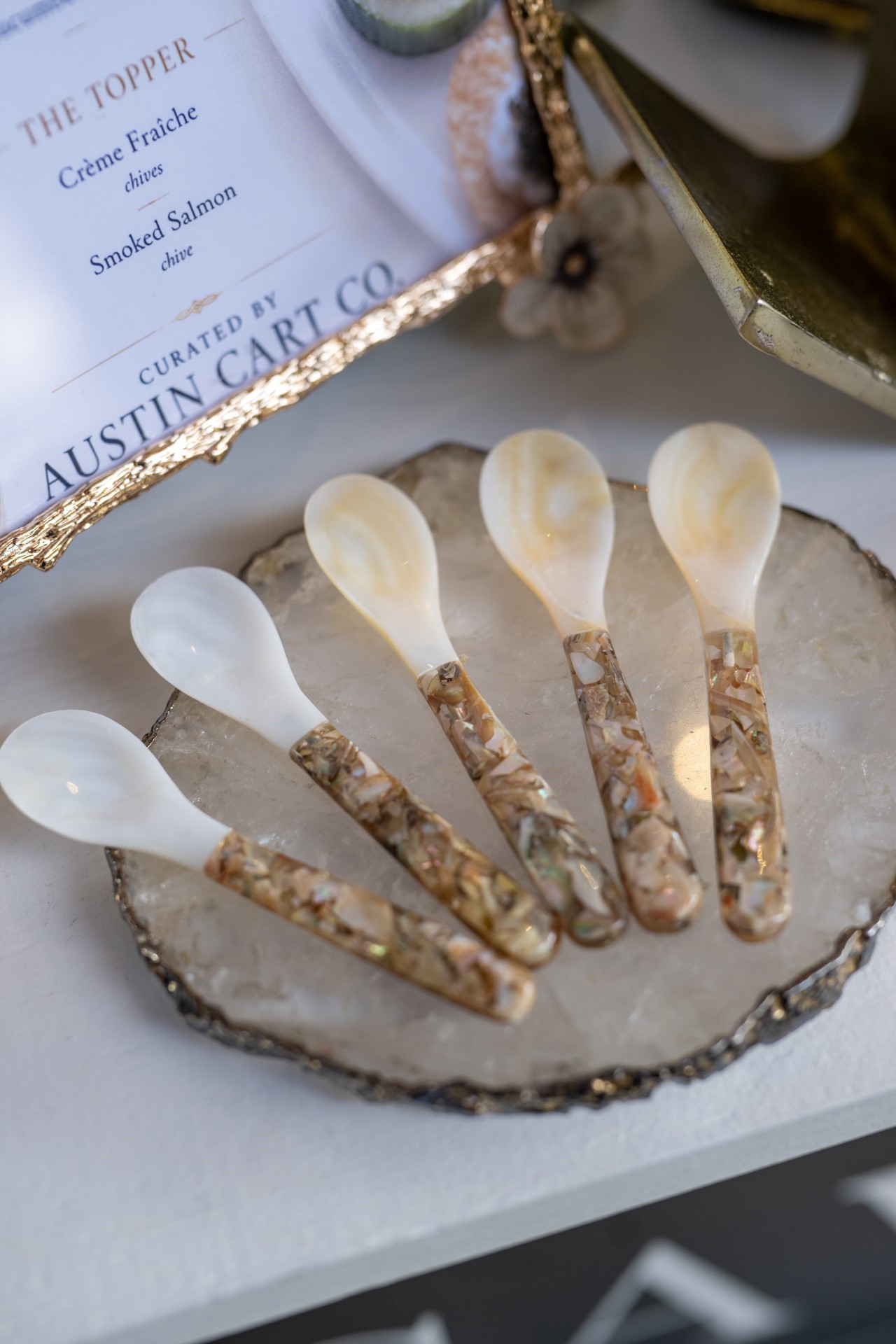 Six small spoons with white bowls and gold handles arranged on a decorative dish, with a framed certificate in the background.