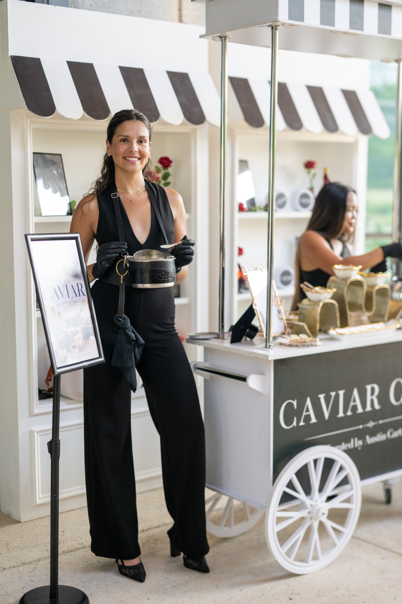 Woman standing at caviar stand with striped awning, smiling, holding a small container, with a display of caviar jars behind her.